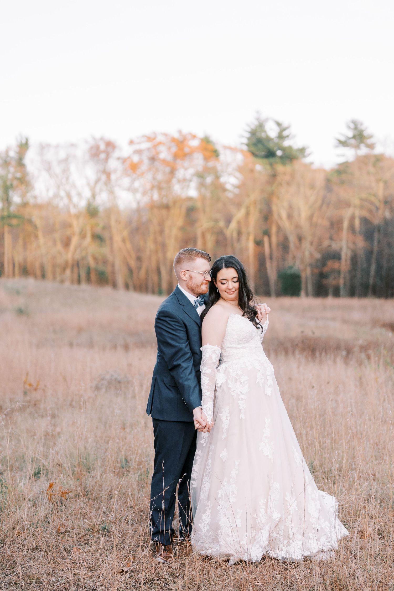 A couple stands together in a rustic autumn field wearing wedding attire with bare trees in the background.