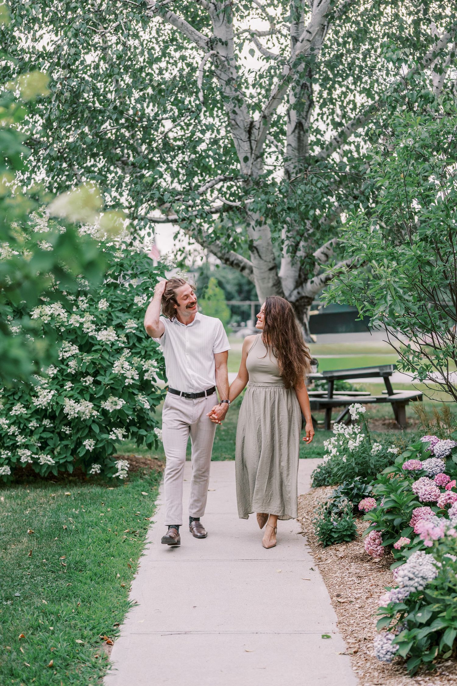 A couple strolls together along a garden path lined with pink hydrangeas and lush greenery.