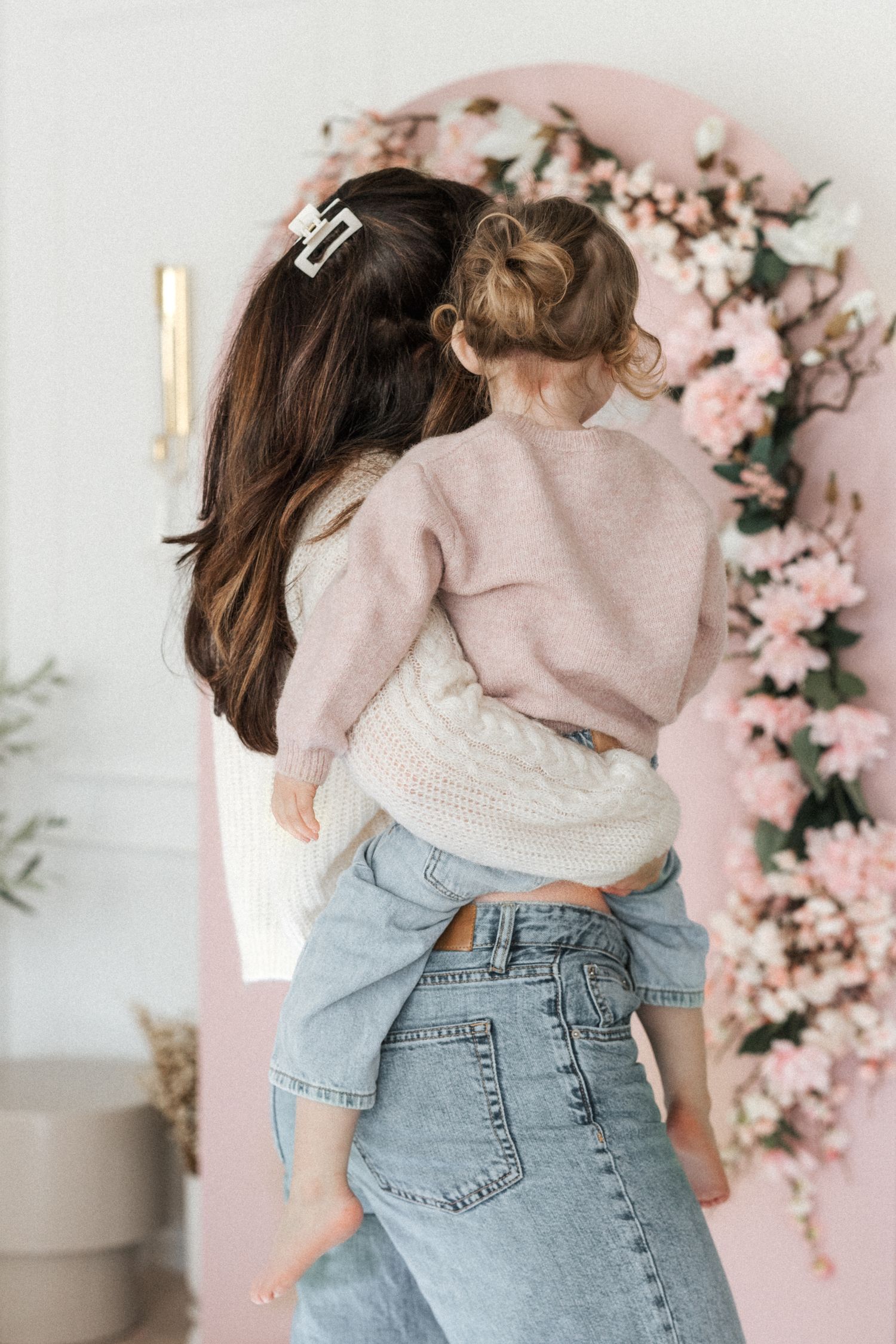 Mother with dark hair holding toddler in front of pink floral arch decoration indoors.