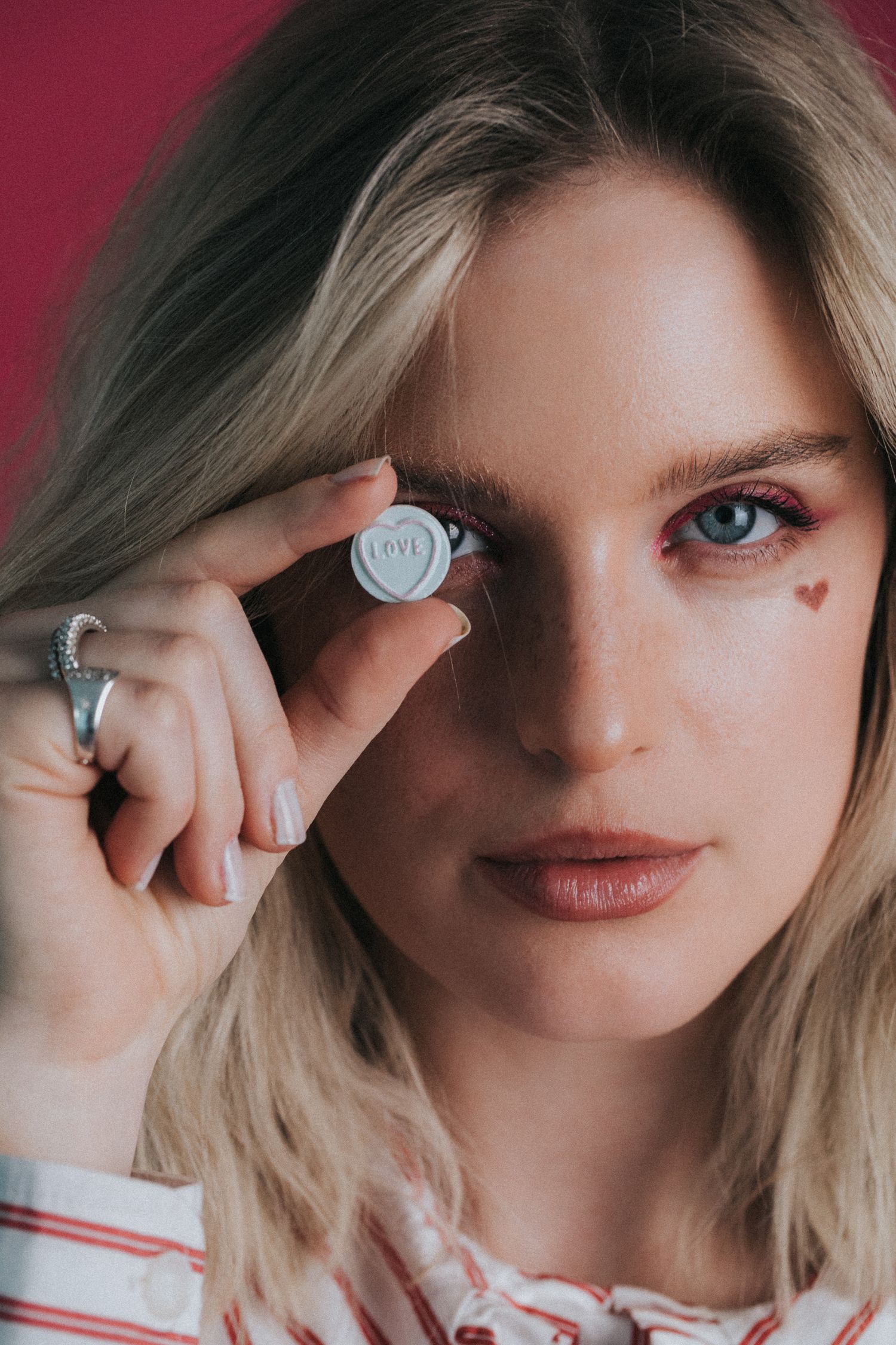 Blonde woman holding silver love coin near eye with small heart drawn on cheek.