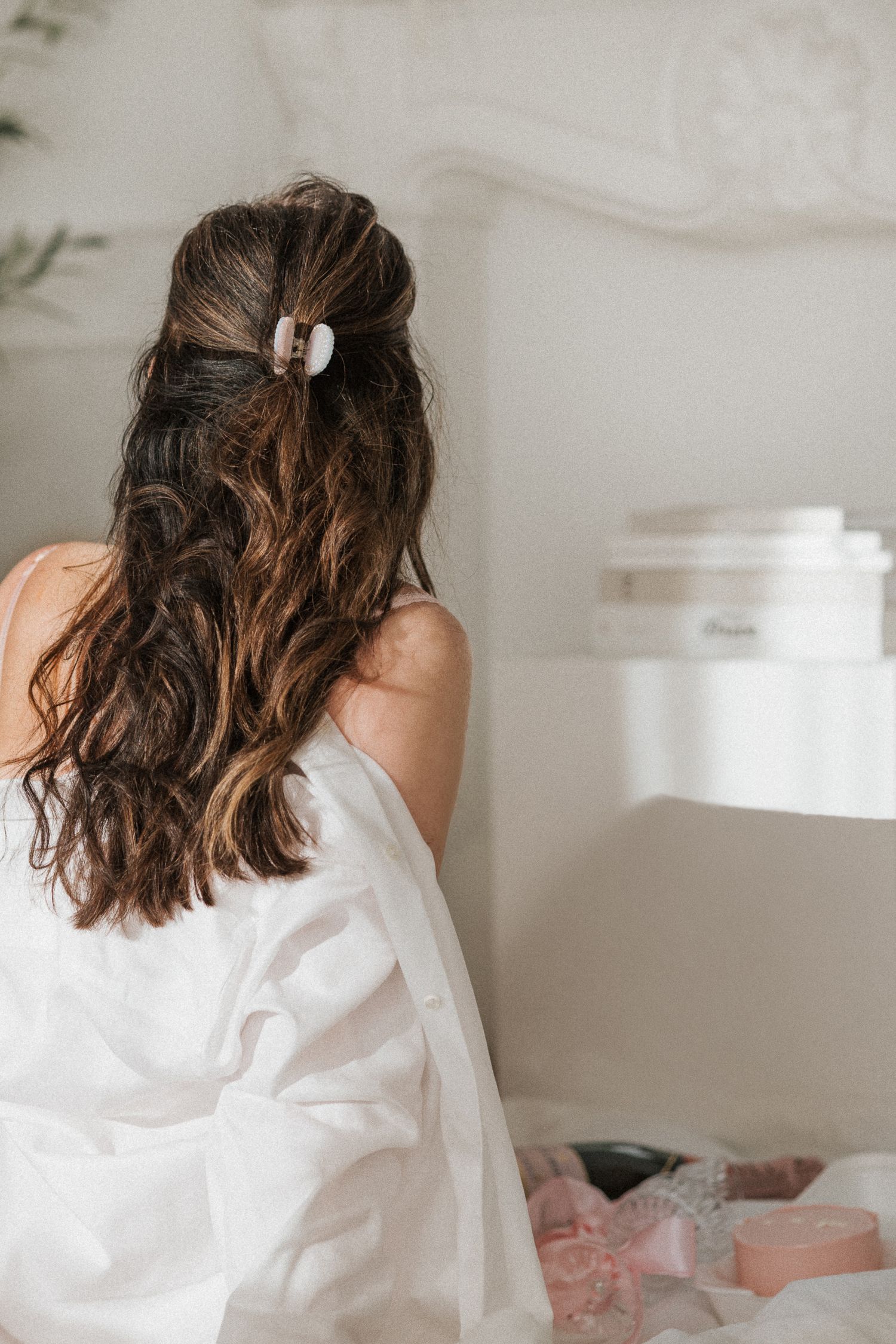 Back view of woman with long brunette wavy hair styled with claw clip in white bedroom setting.