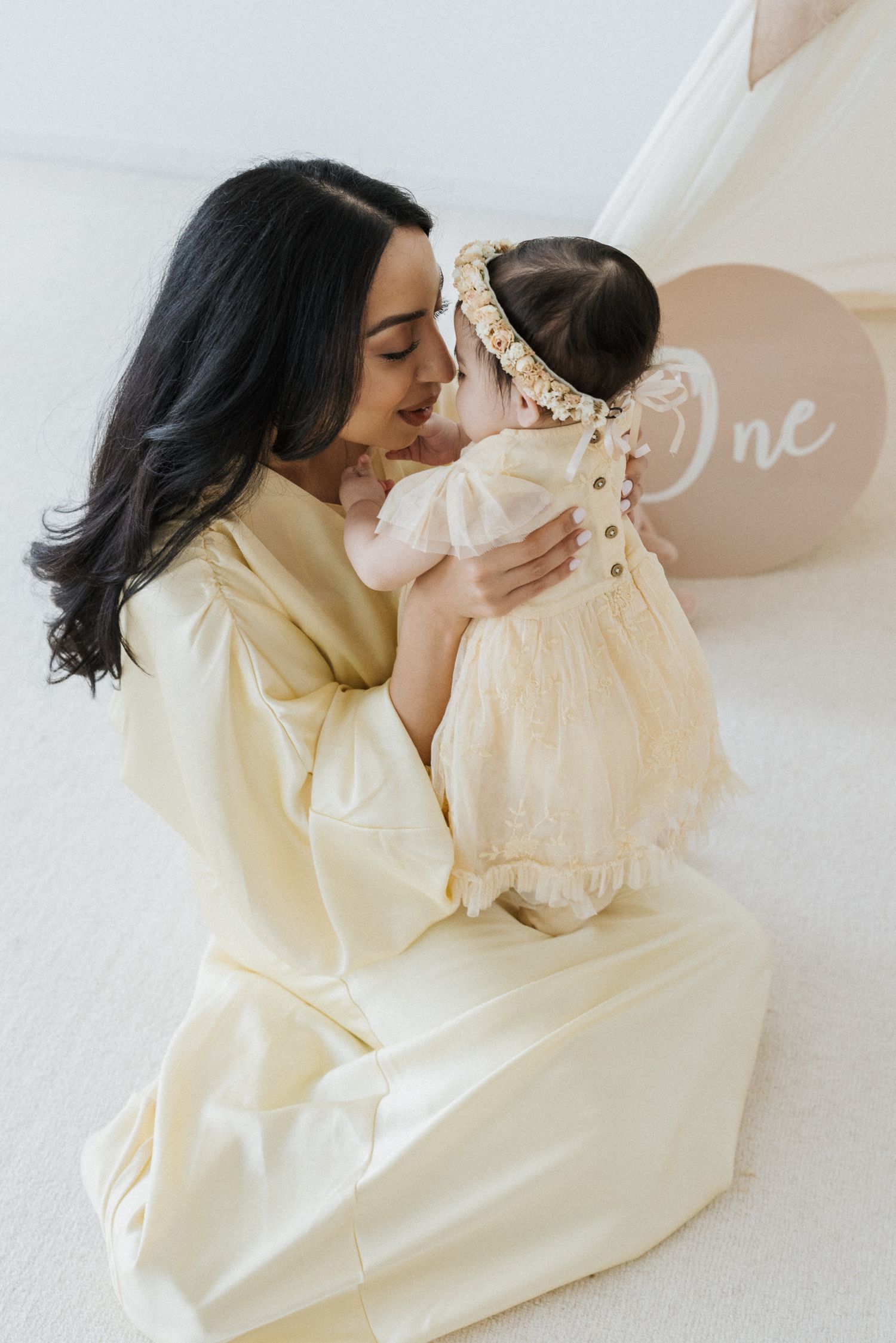 Dark-haired mother in yellow dress kissing baby in cream dress at first birthday celebration.