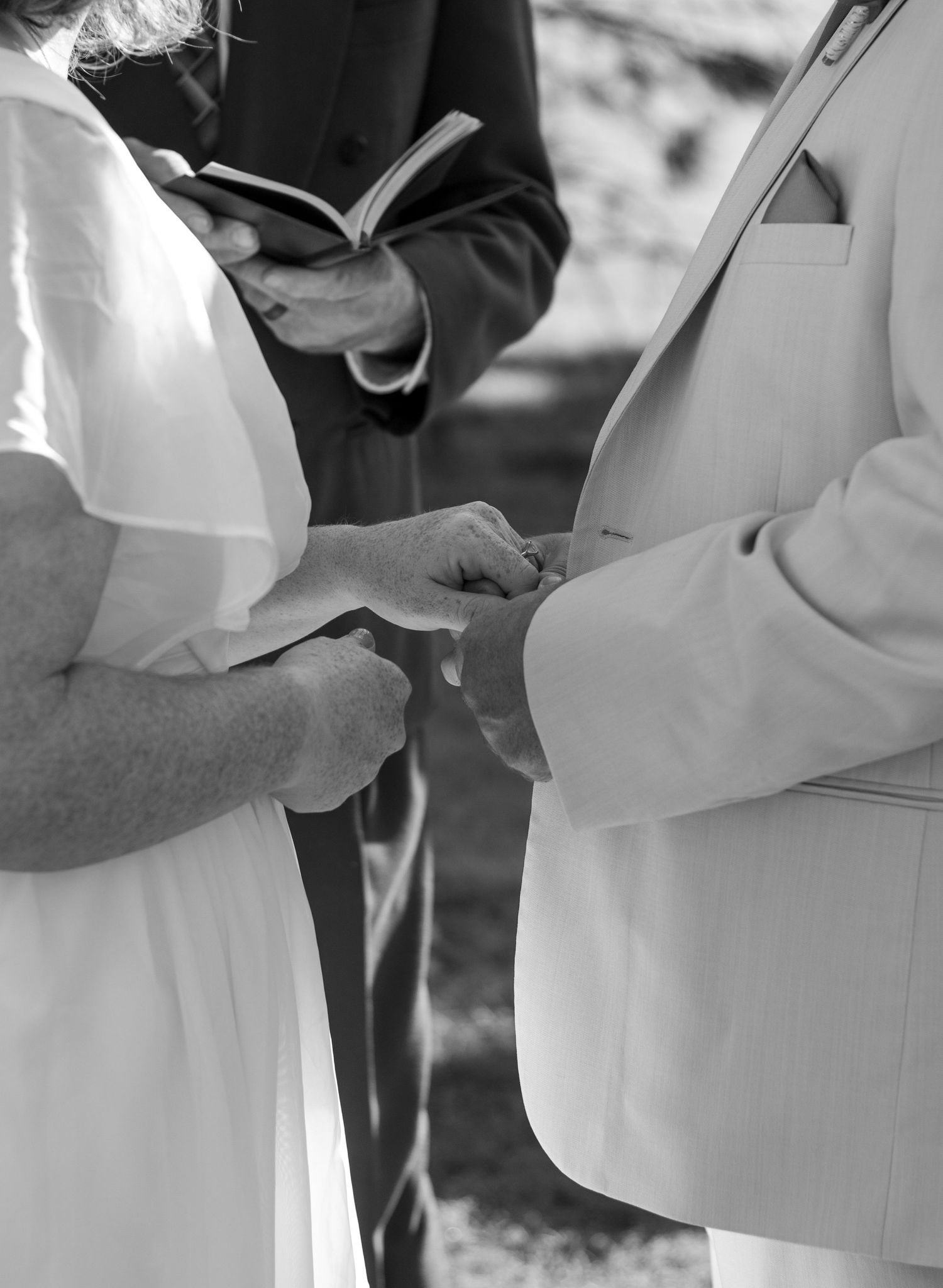 Close-up view of hands holding hands placing the rings during an outdoor wedding ceremony.