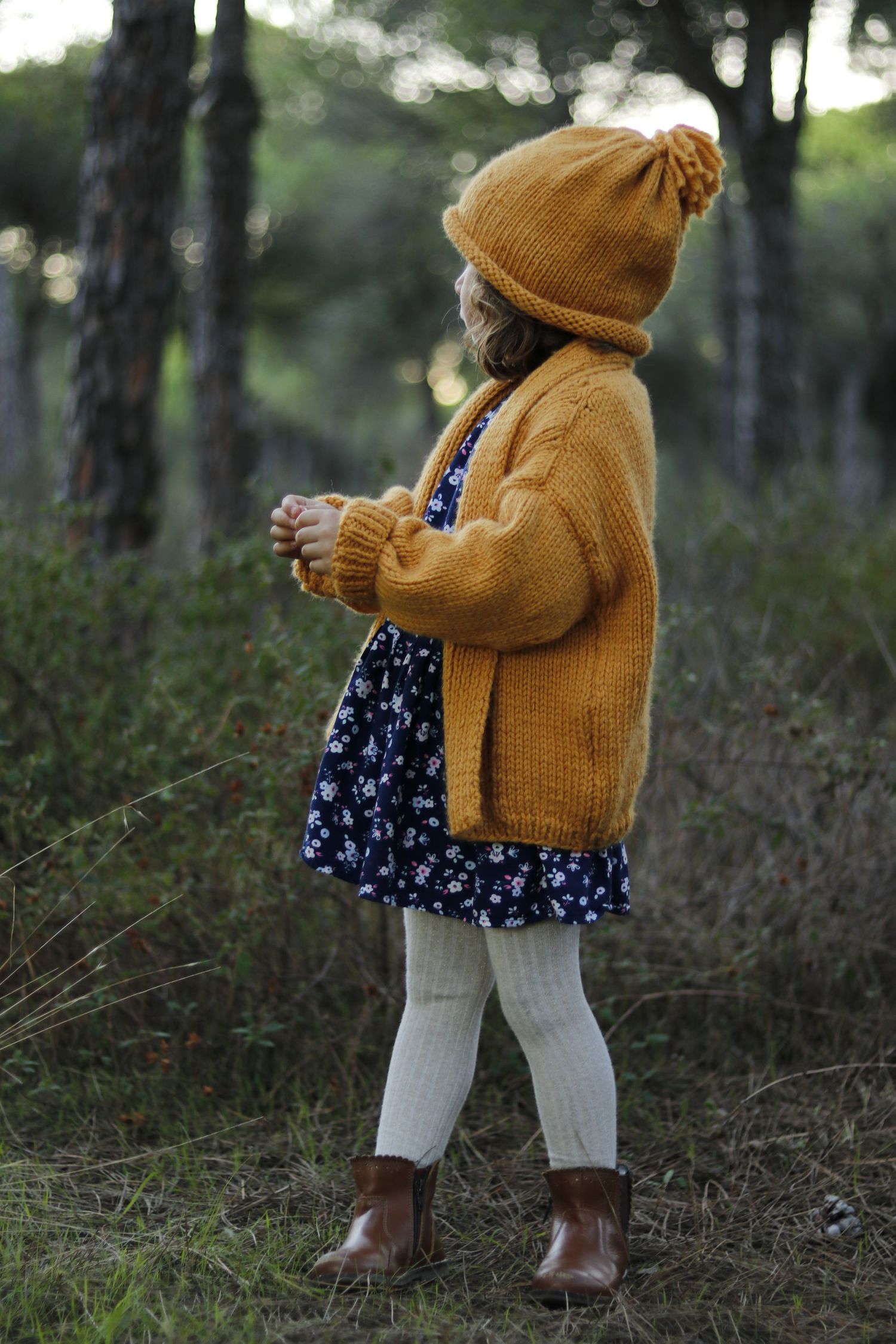 Small figure in woods wearing mustard cardigan, floral dress, and beanie with knit leggings.