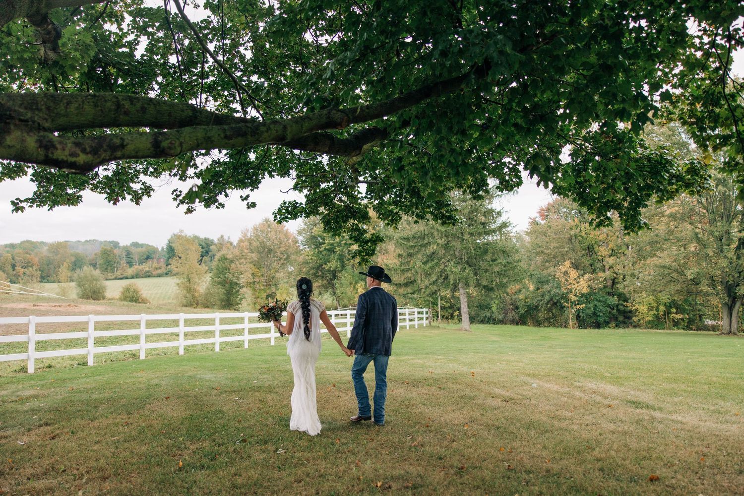 Picturesque outdoor wedding ceremony in a green hillside field with white fencing and trees in the background.