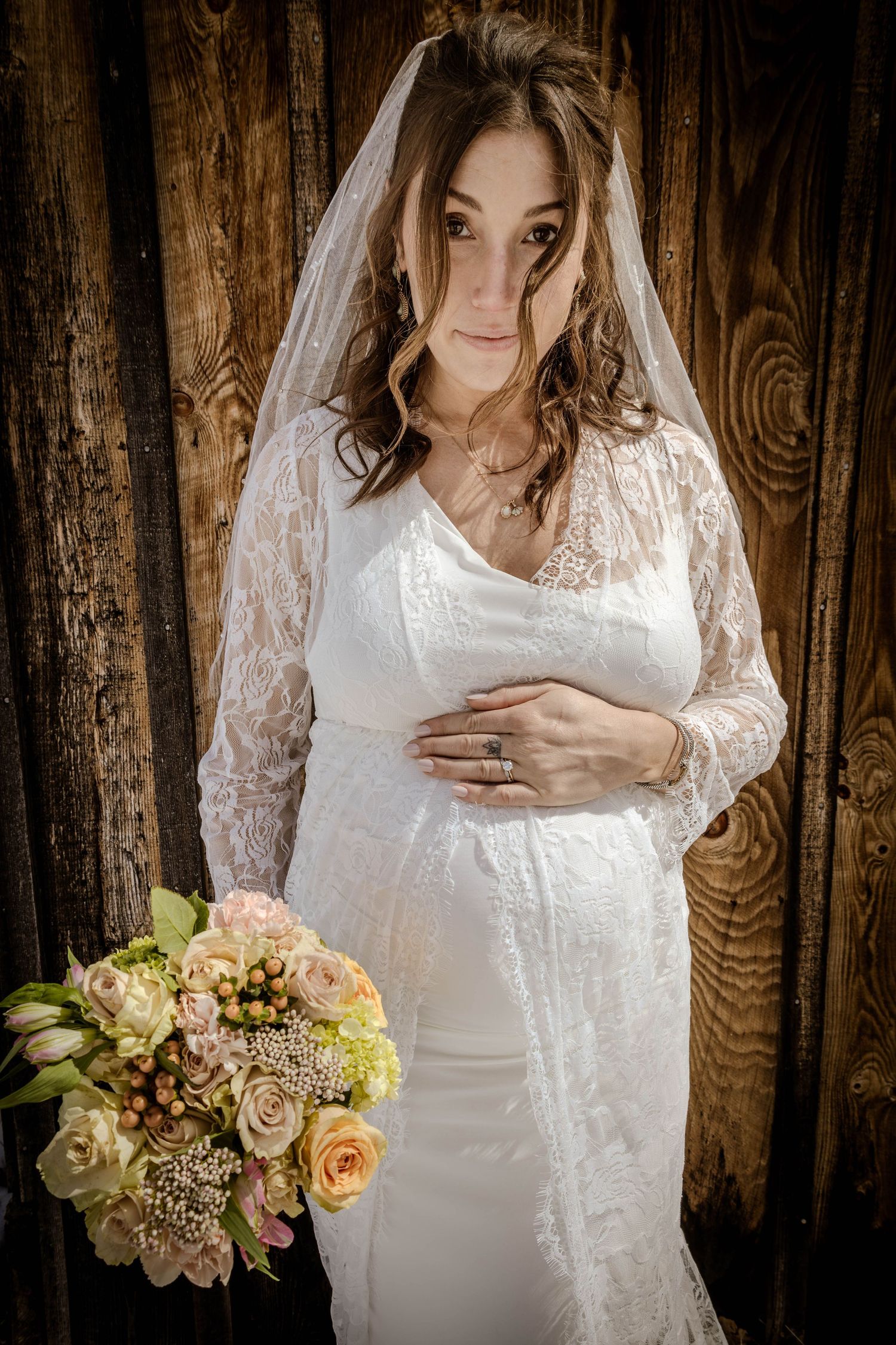 Bride holding pregnant belly and flowers in front of a barn.