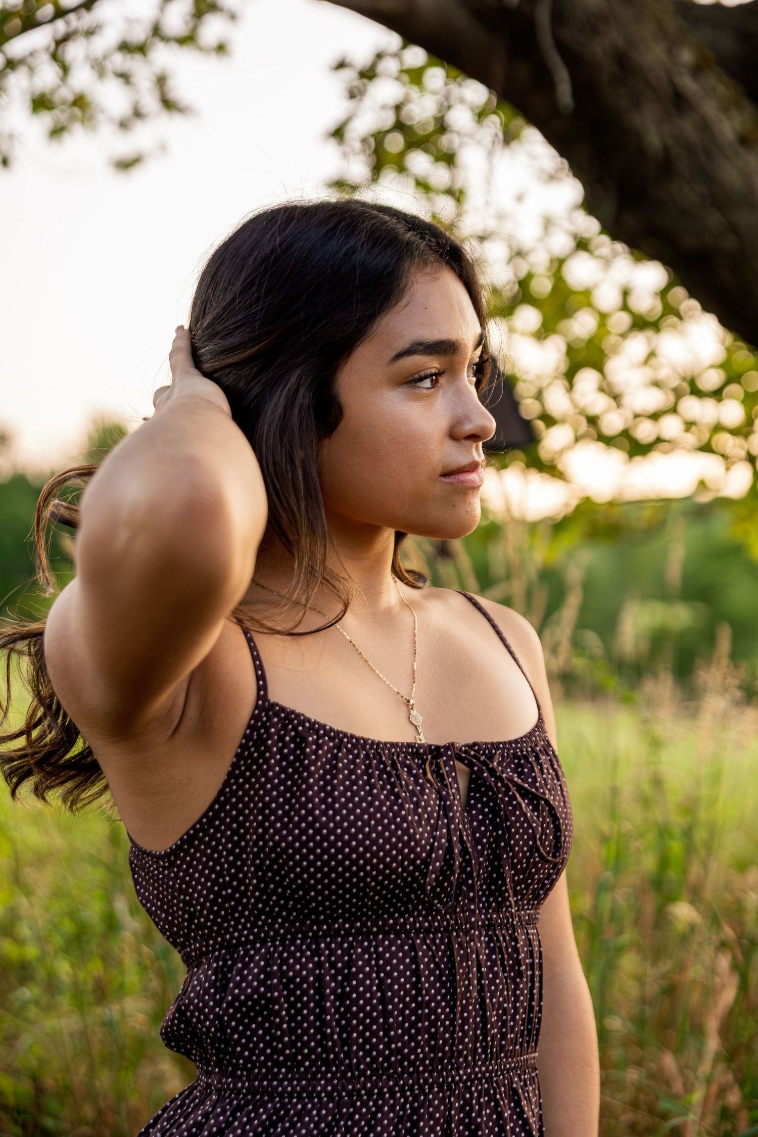 Young woman in polka dot sundress poses outdoors at golden hour, hand in dark hair, with lush green trees behind her.