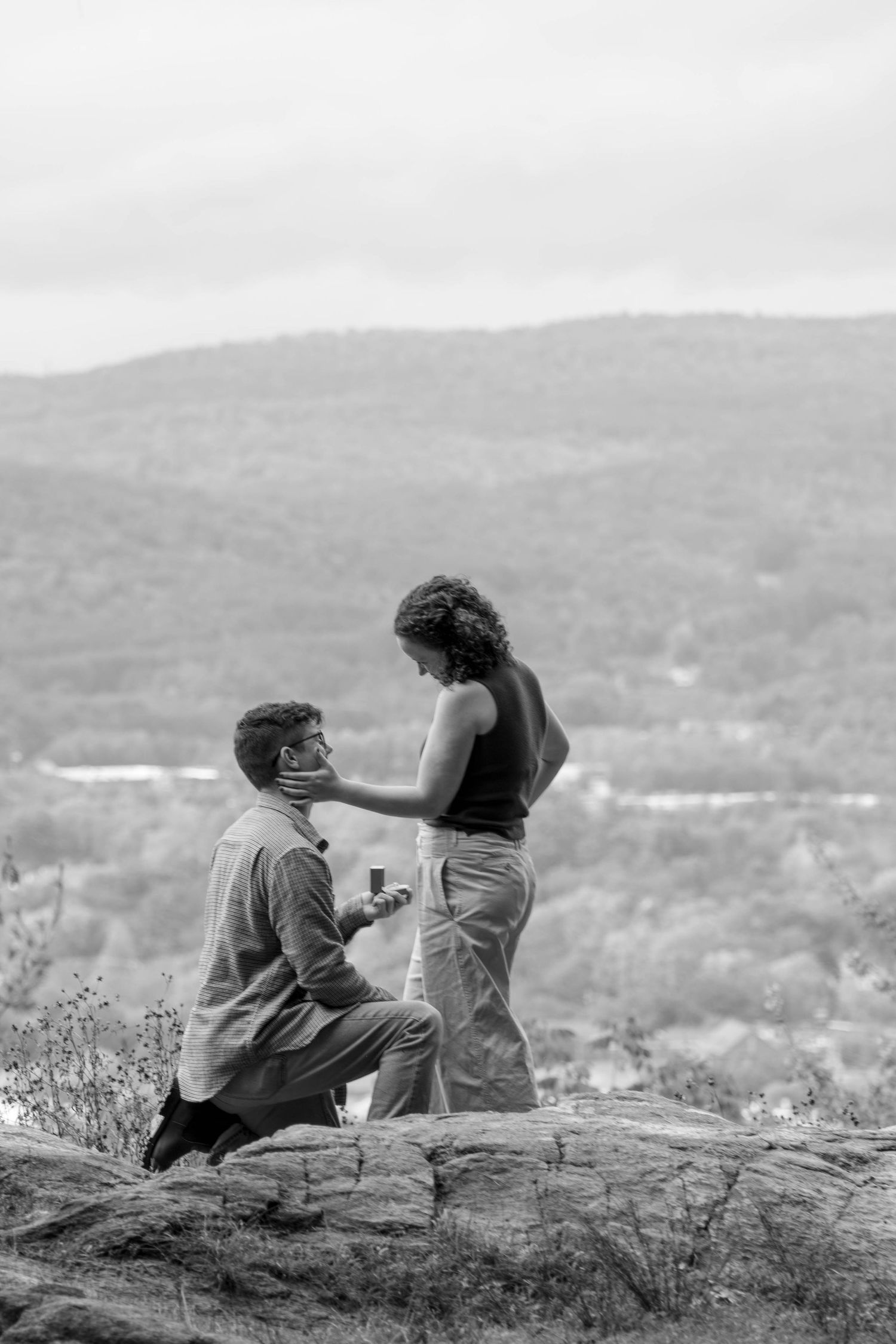 Black and white photo of couple sitting on rocky overlook with mountain vista in background.