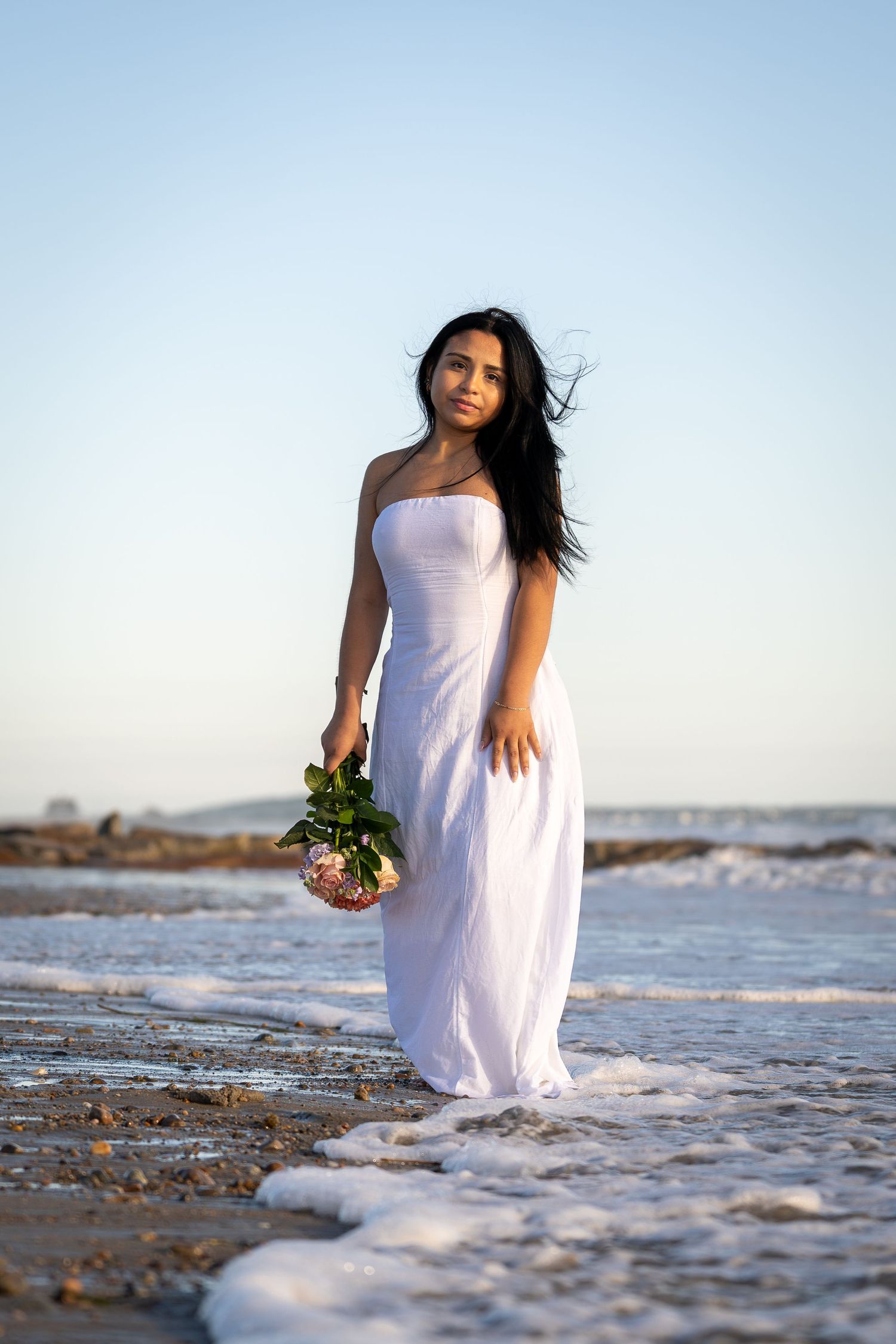 Woman in flowing white dress holds bouquet while standing on beach at sunset.