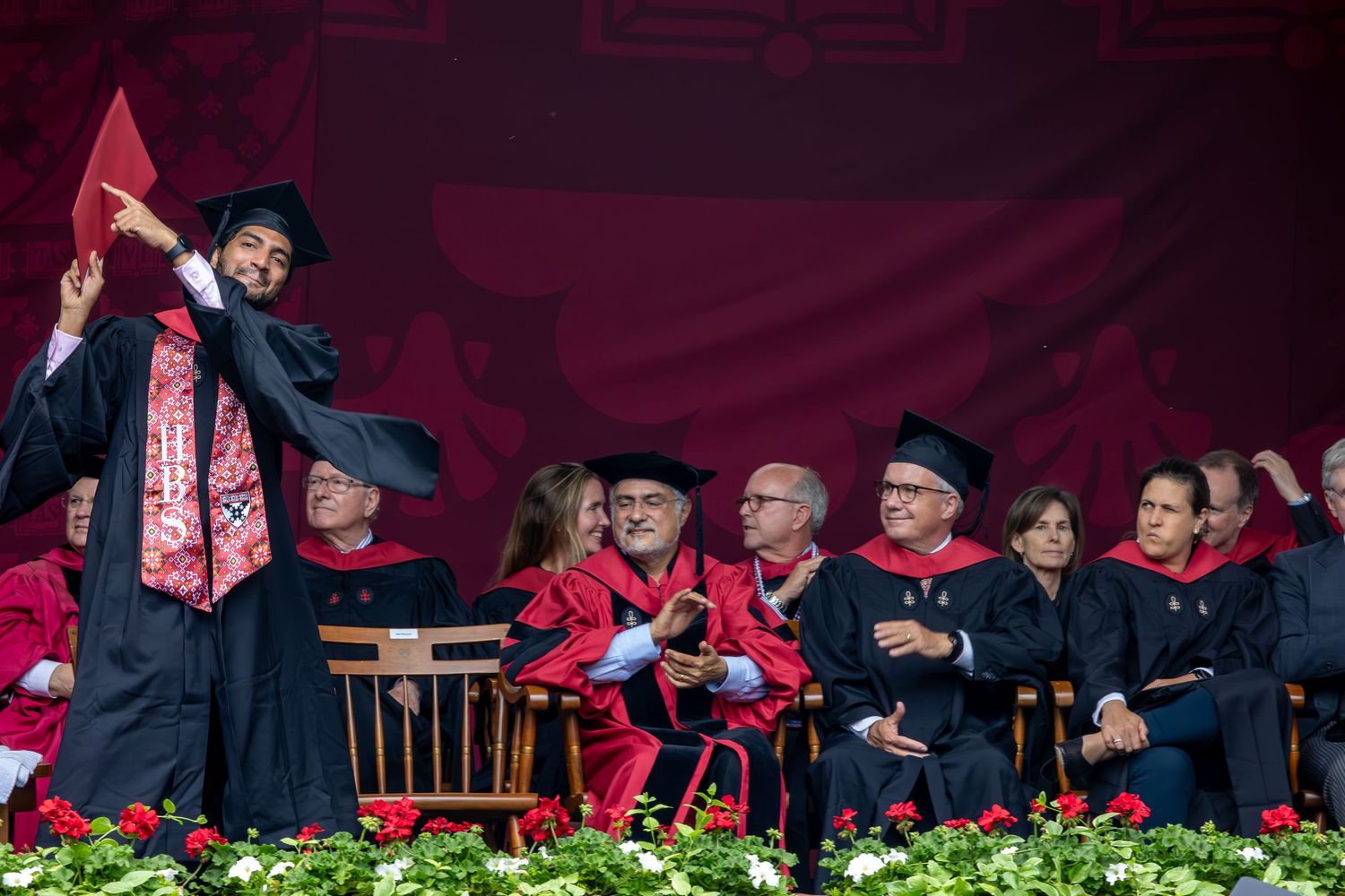 Graduate celebrates with raised diploma during outdoor commencement ceremony with faculty in background.