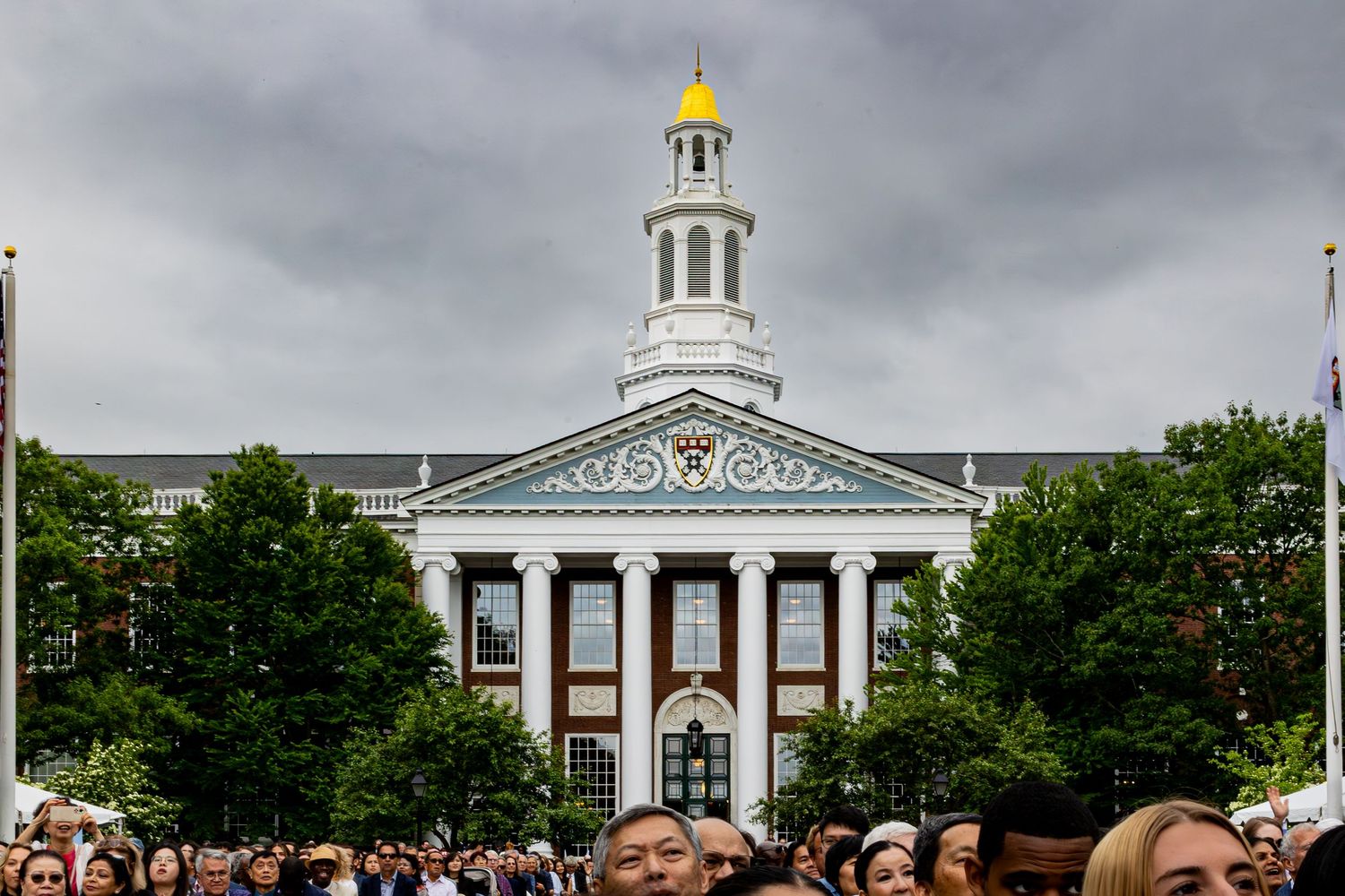 Crowd gathered in front of Harvard Business School's Baker Library with white columns and gold dome under cloudy skies.