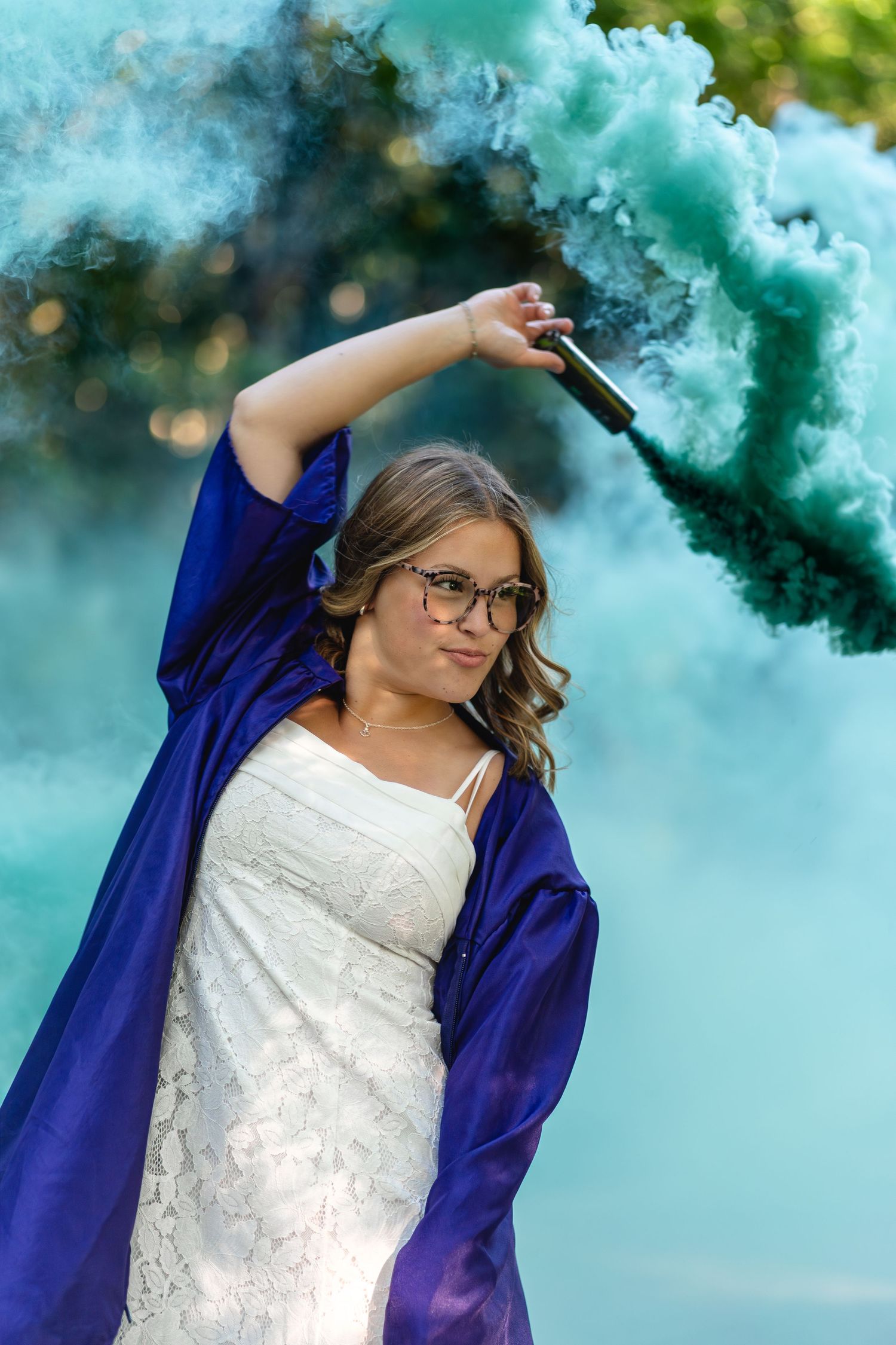Young woman in graduation gown holds teal smoke bomb outdoors.