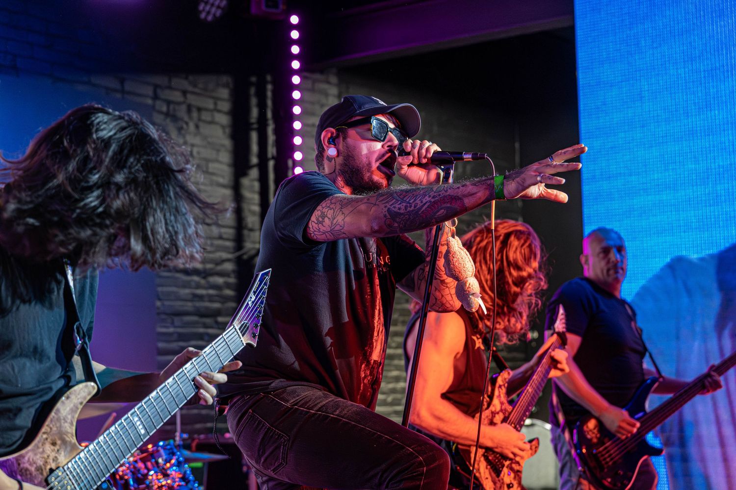 Live rock band performing on stage with dramatic purple lighting, vocalist singing into mic while guitarist plays.