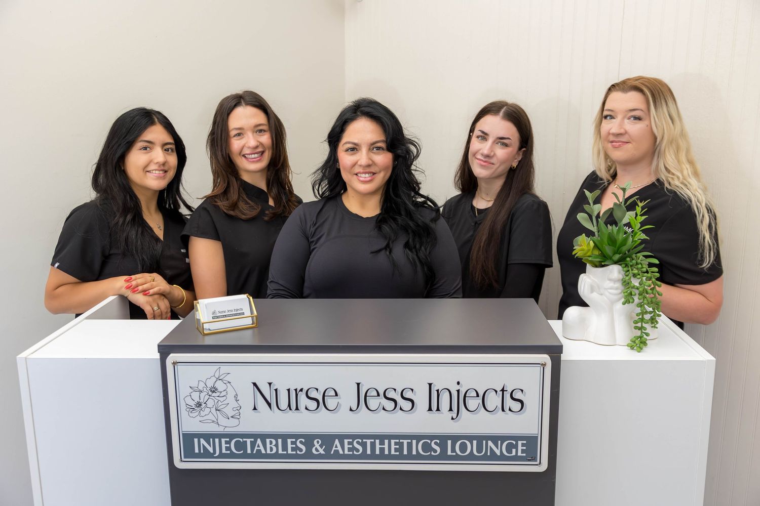 Five women smile behind the Nurse Jess Injects Injectables & Aesthetics Lounge reception desk.