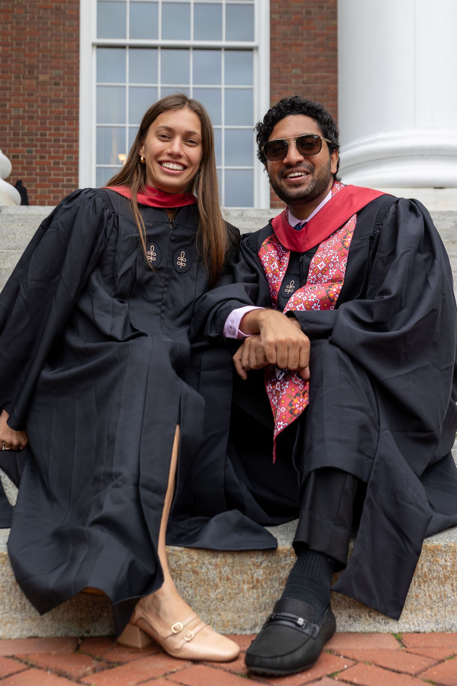 Two graduates in black robes and red stoles sit smiling on steps outside a brick building.