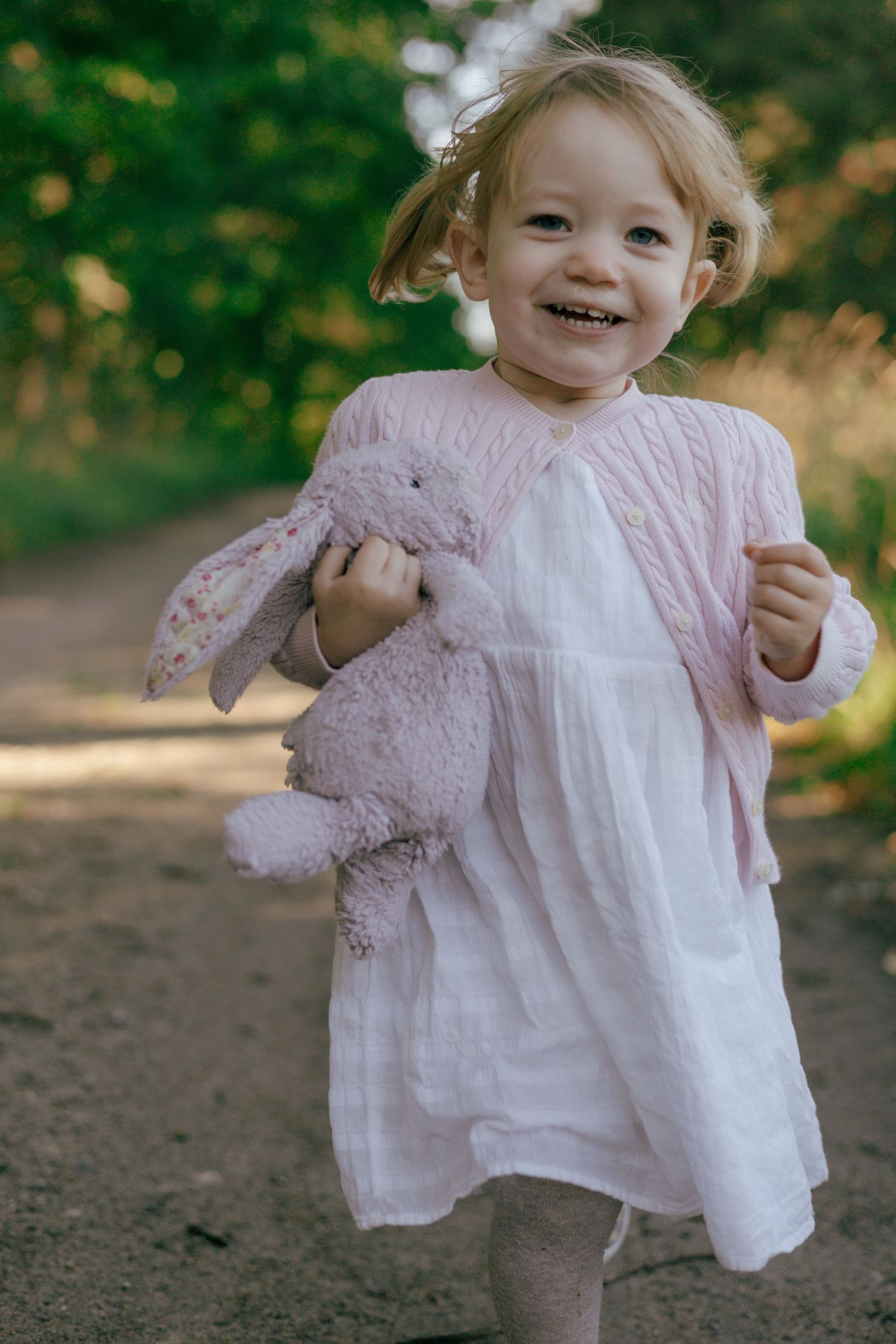 Child in white dress holding pink stuffed bunny while smiling outdoors.