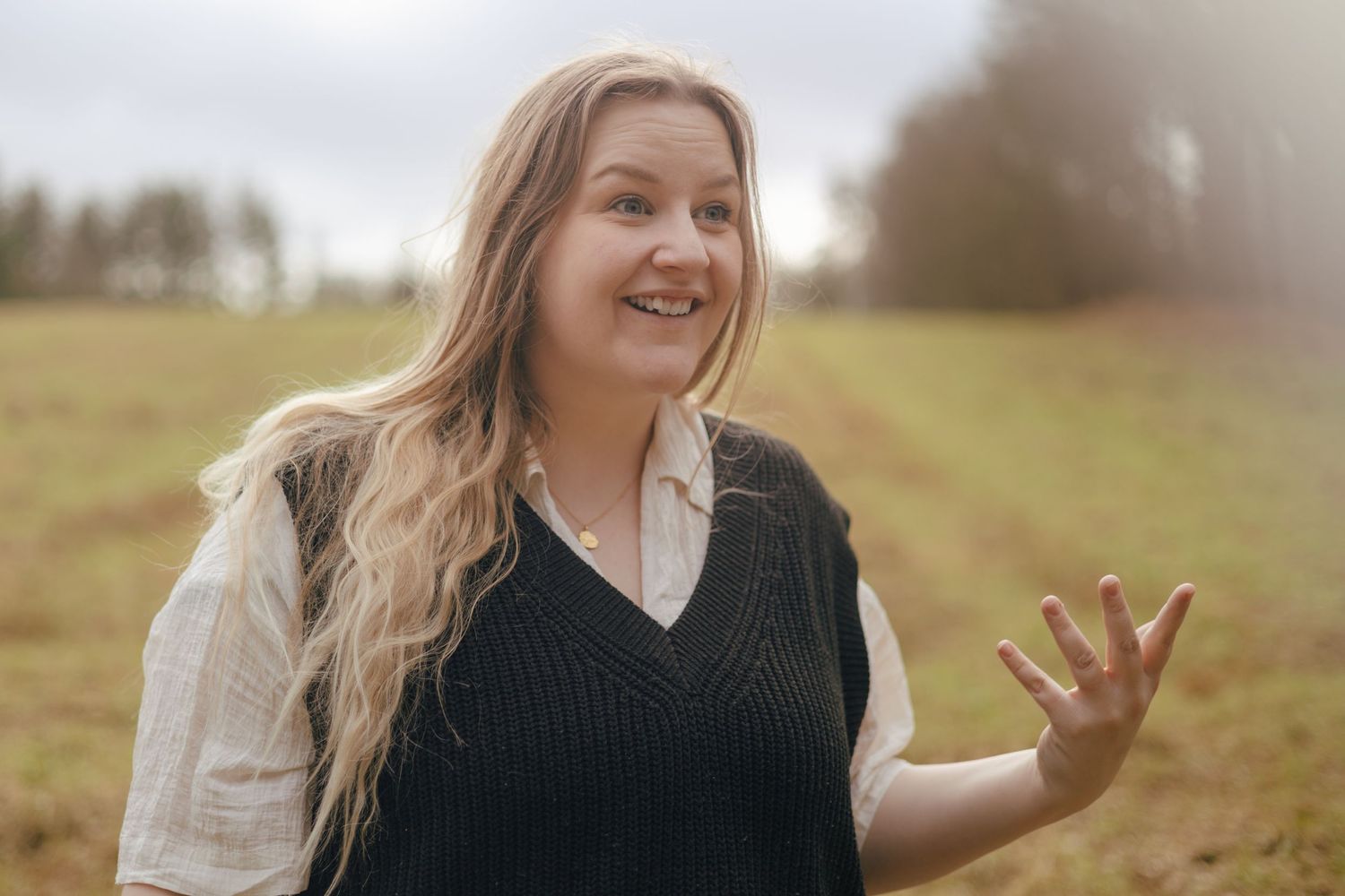 Person wearing black sweater vest and white shirt waves and smiles in a sunlit field on a hazy autumn day.