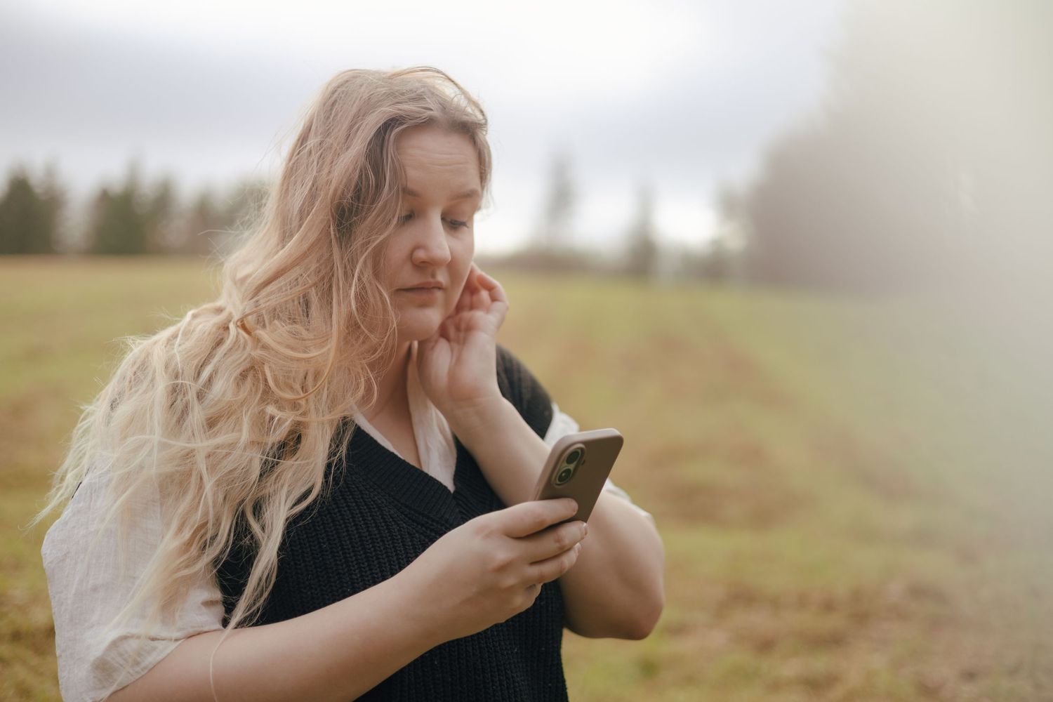 A person with long blonde hair stands in a field looking at their phone on an overcast day.
