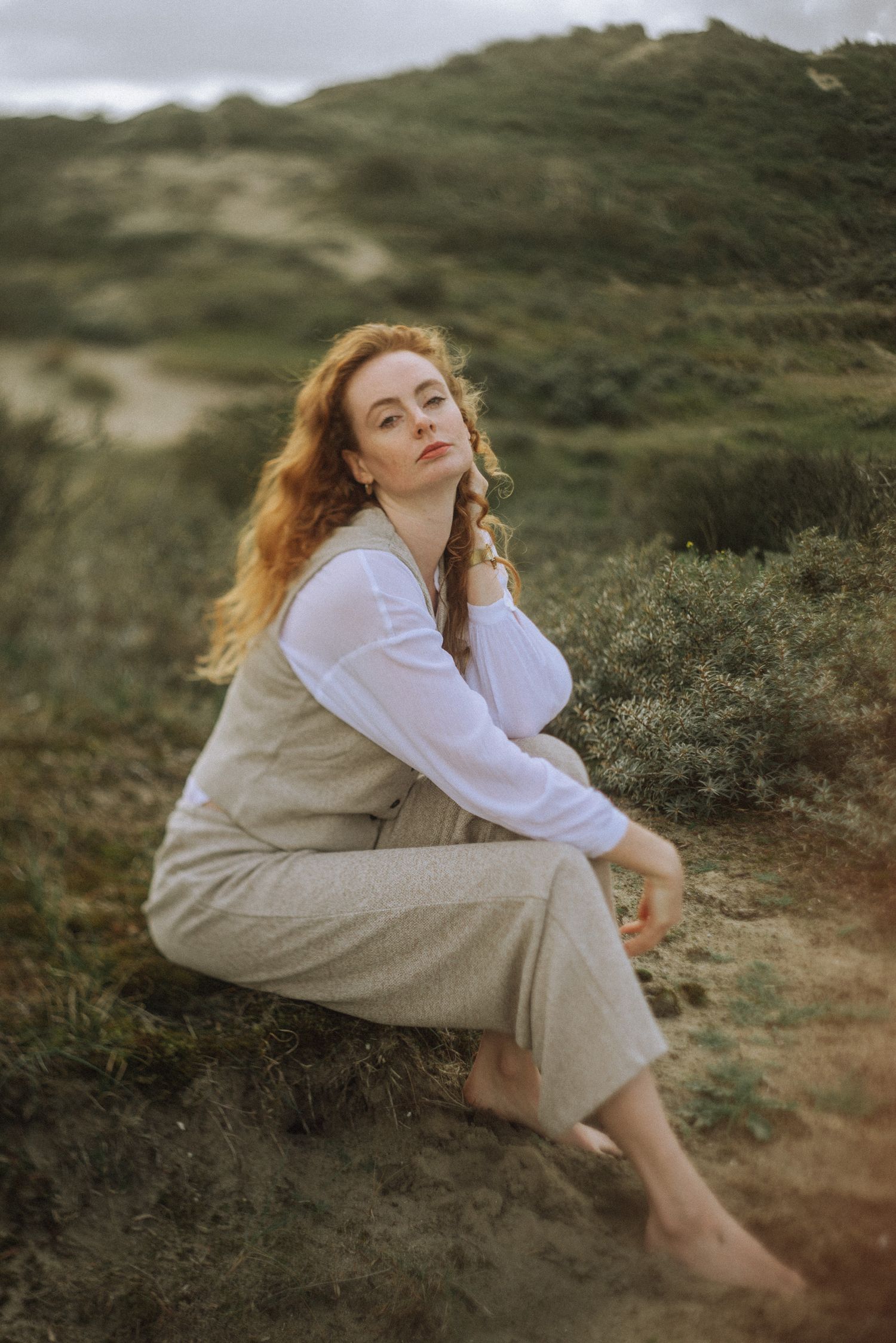 Figure in white and beige clothing sitting contemplatively on sandy hillside during golden hour.
