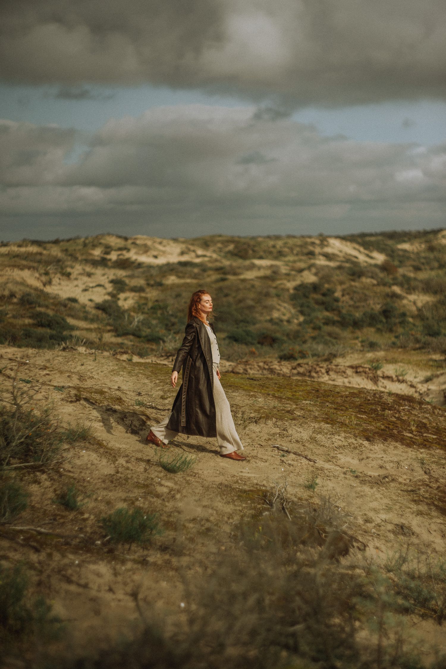 Person in casual attire walking along rugged coastal dunes under dramatic cloudy skies.