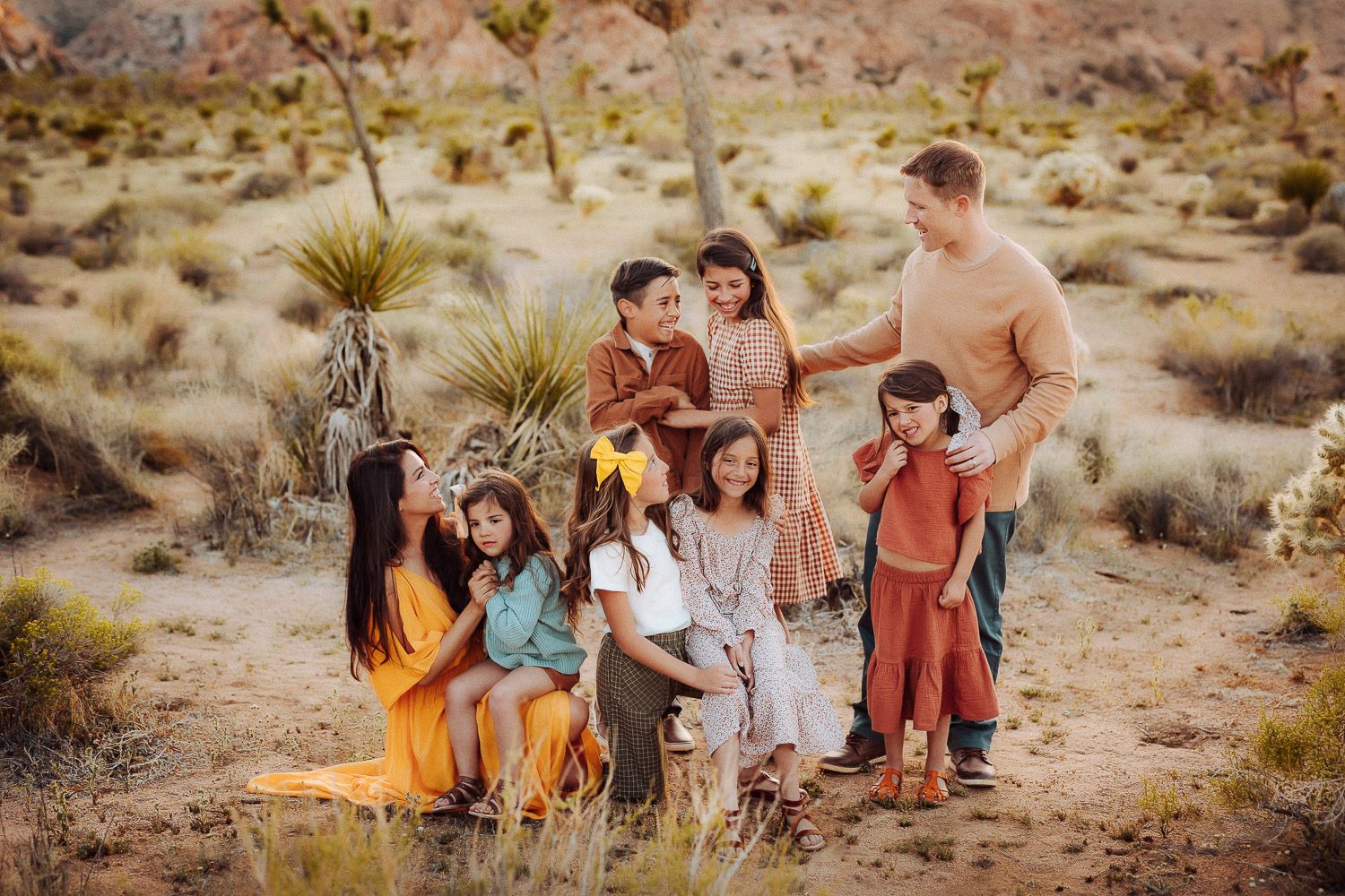 A joyful family of eight embracing in the desert at sunset, wearing coordinated earth-toned outfits with mountains in th