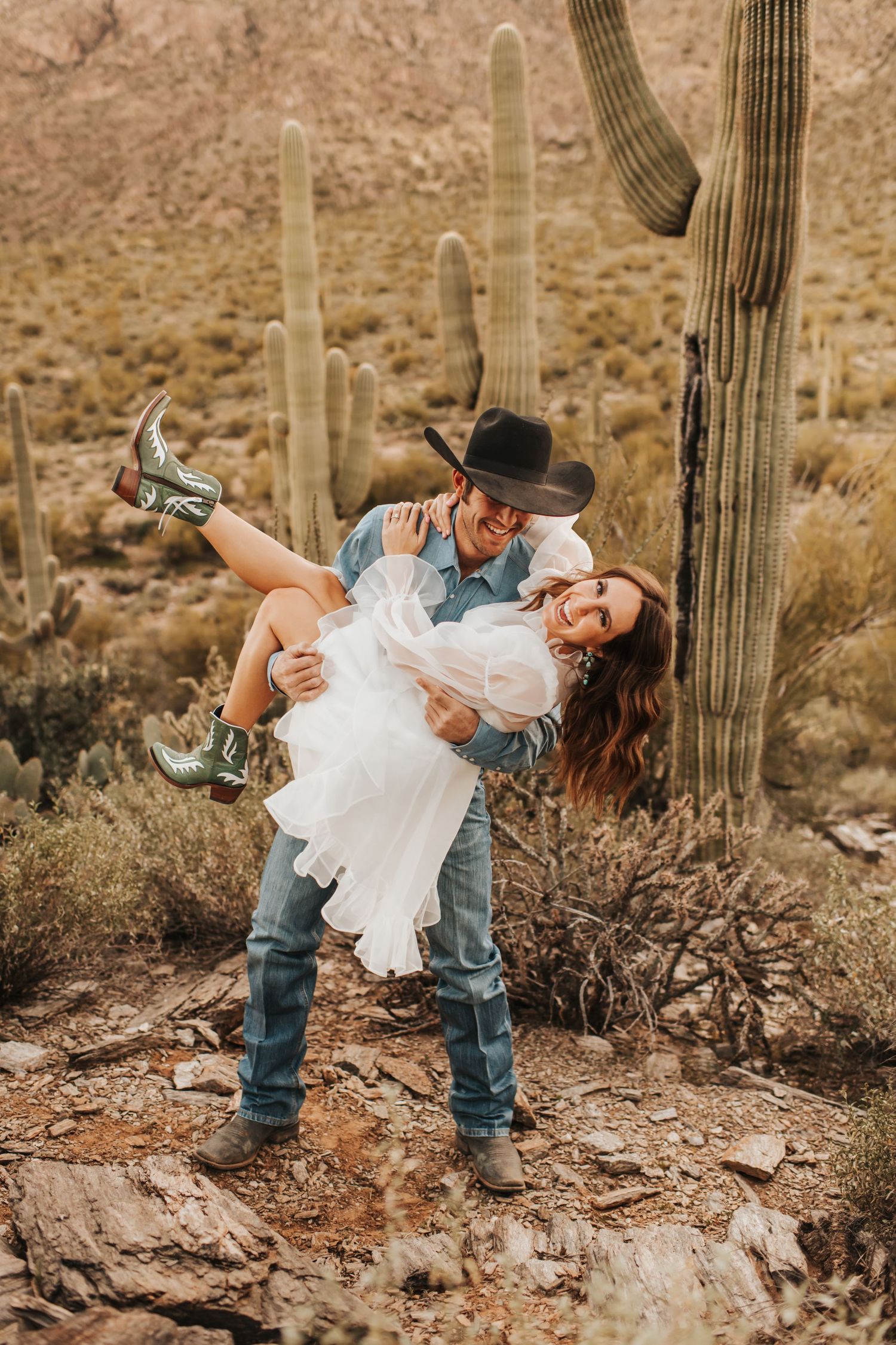Man in a cowboy hat dipping his fiance wearing cowgirl boots in front of a cactus