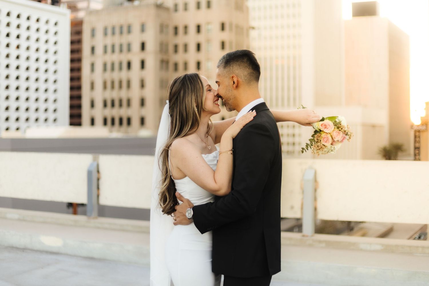 A newly married couple lean in for a kiss. The bride has her bouquet behind her husband's back.