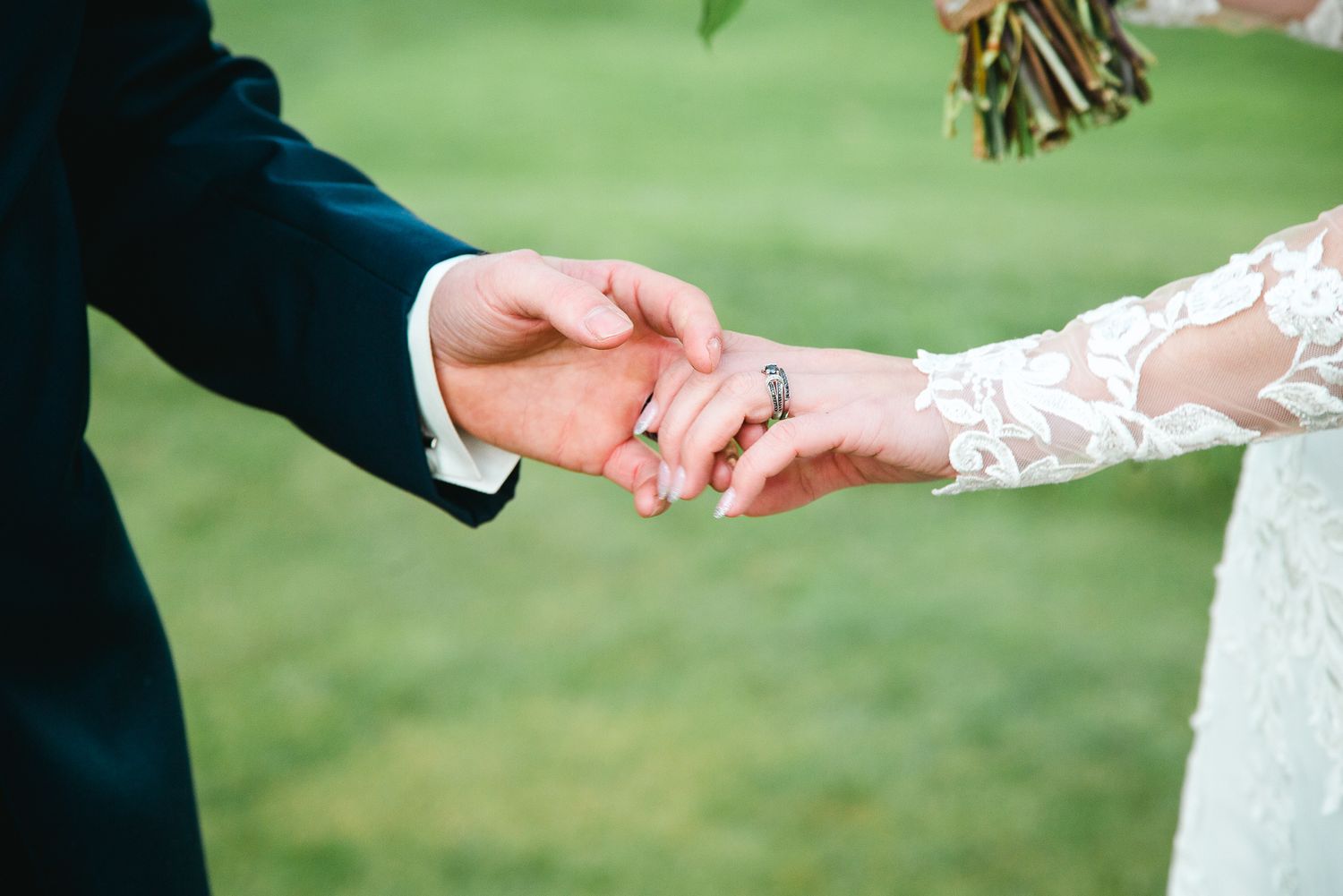 Close-up of newlywed couple holding hands outdoors, with bride wearing lace sleeve and groom in dark suit.
