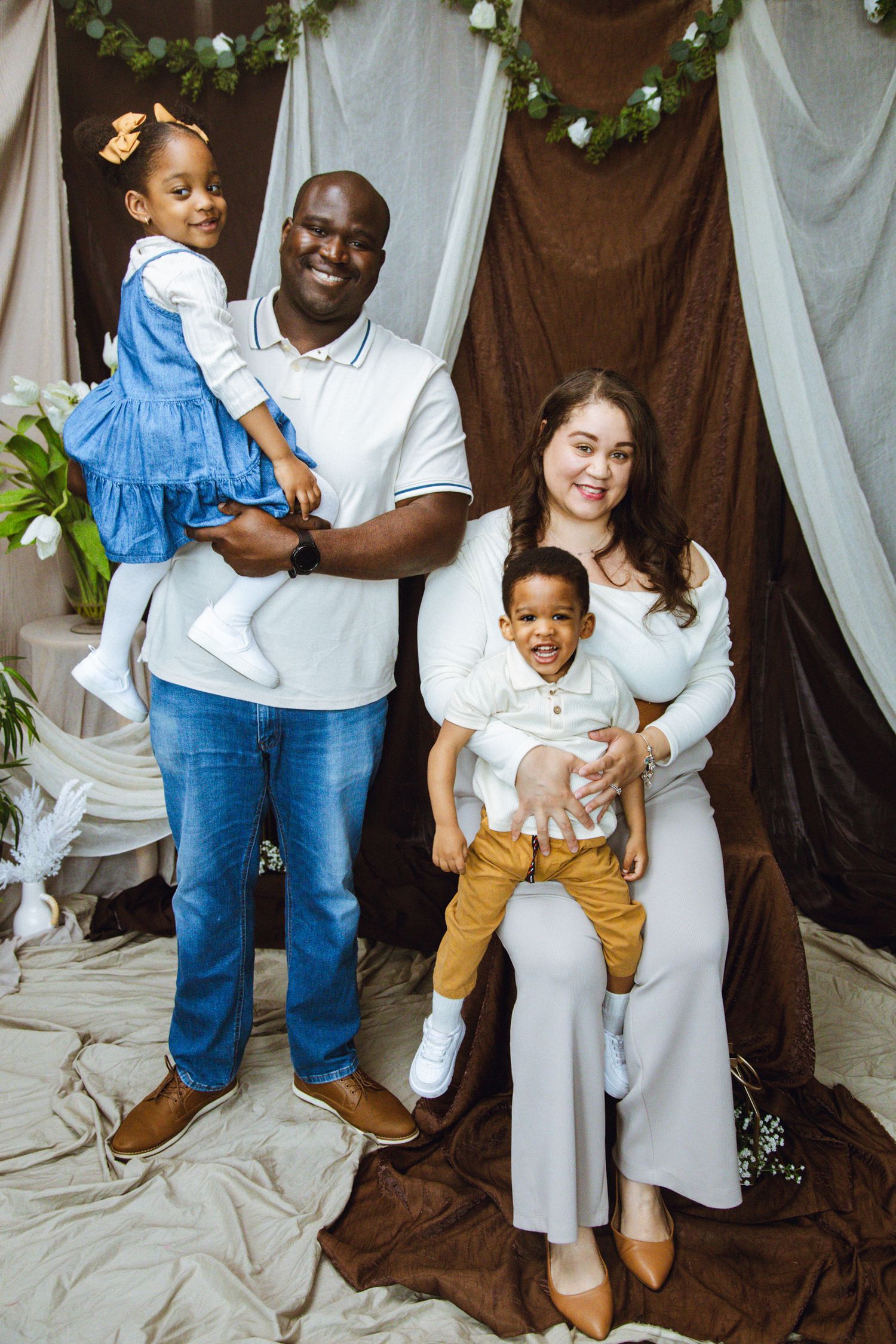 Happy interracial family of four posing in a decorated studio, parents holding their two young children, smiling warmly.