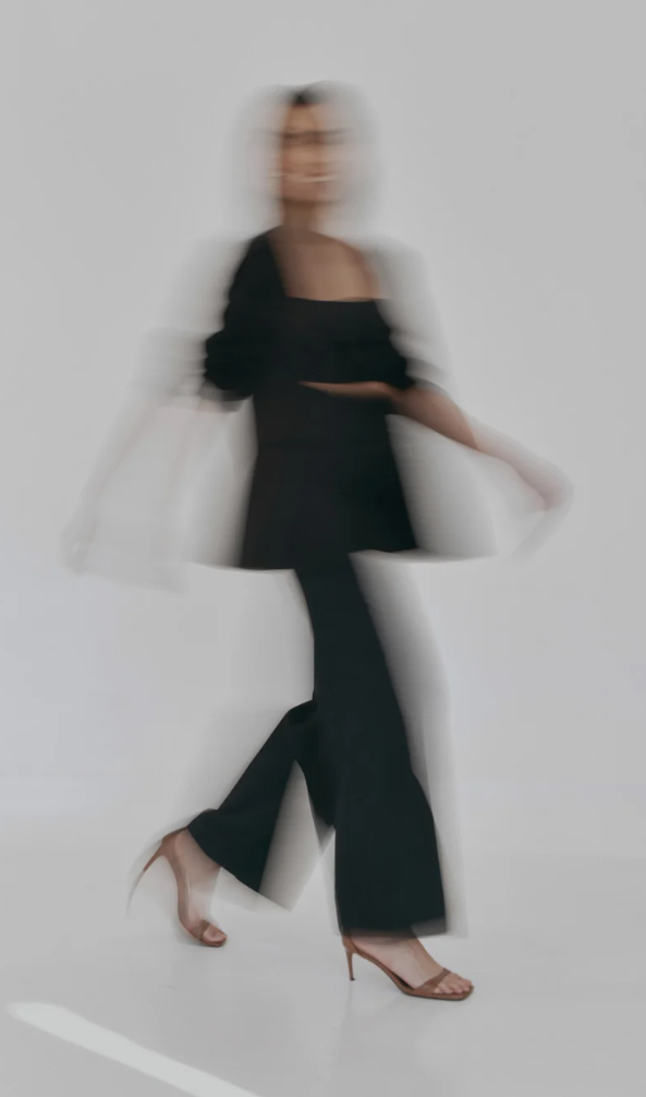 Motion blur captures a figure in black attire and heels dancing against a white background.