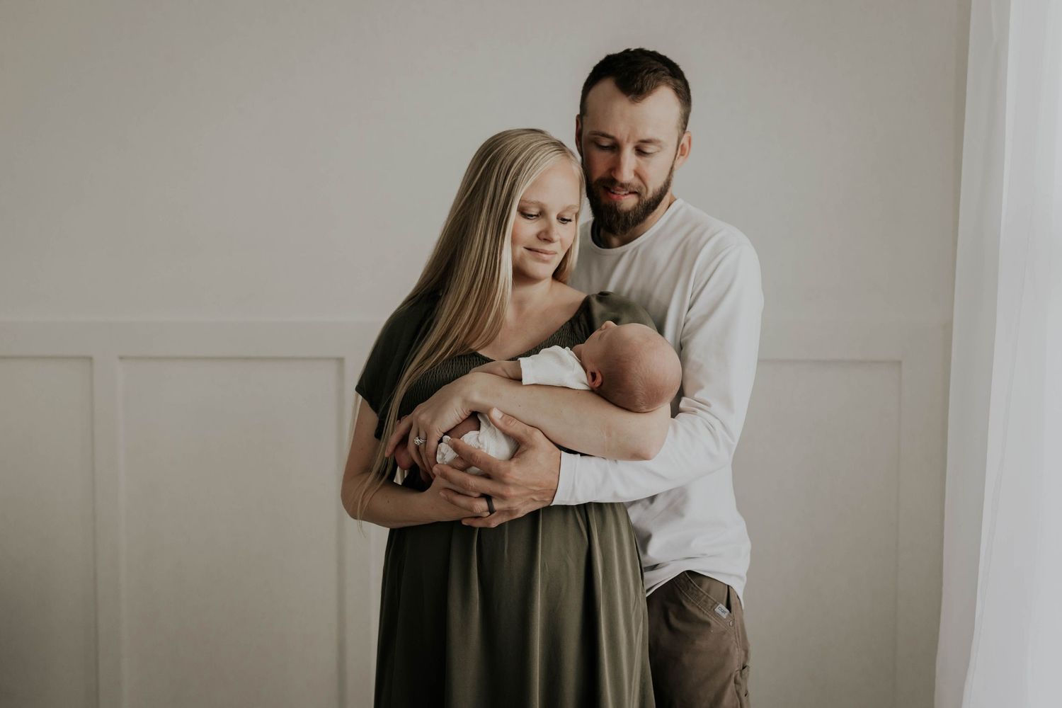 A couple tenderly holds their newborn baby together, standing against a soft white wall in a warm family portrait.