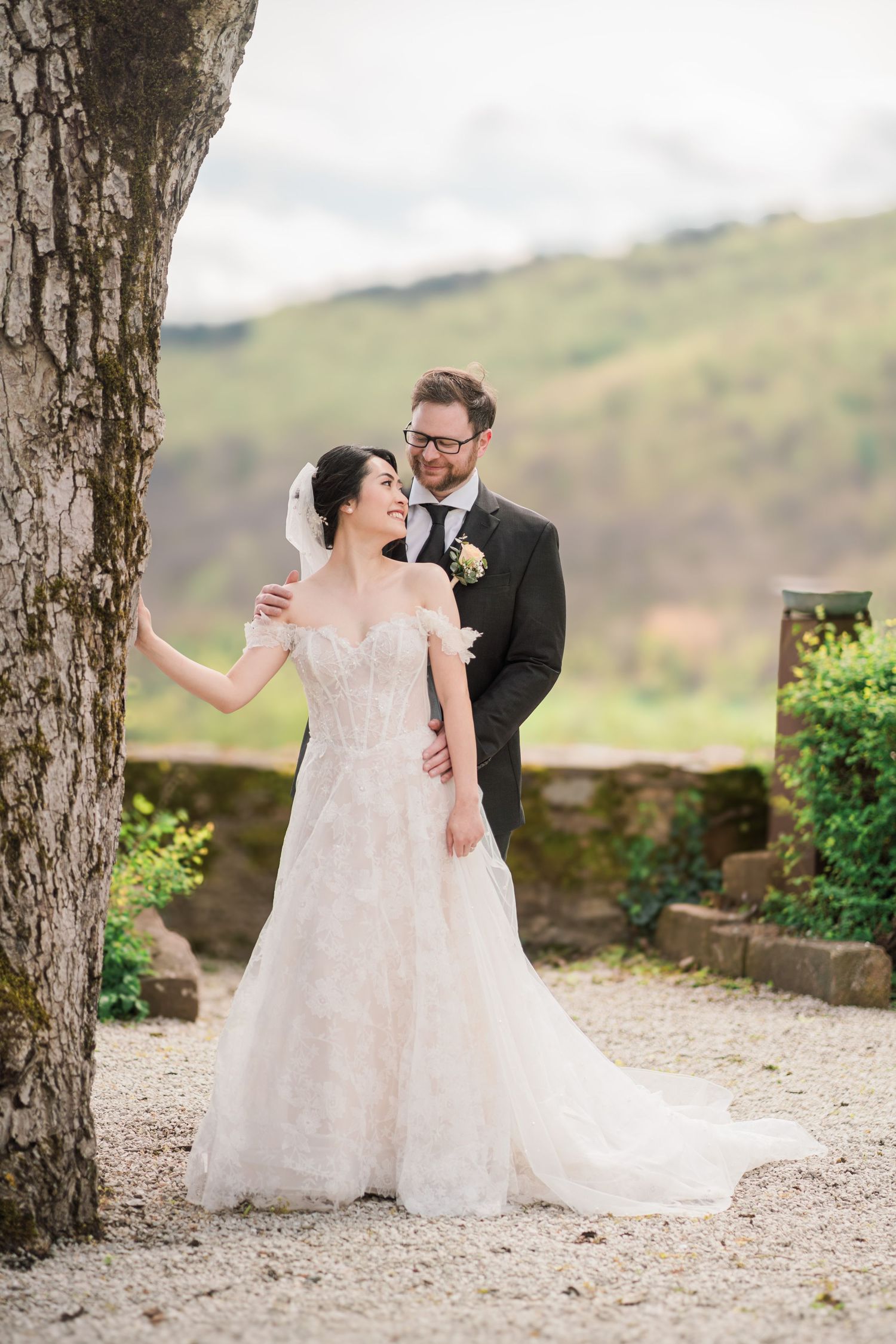 Romantic outdoor wedding portrait under large tree with scenic mountain backdrop.