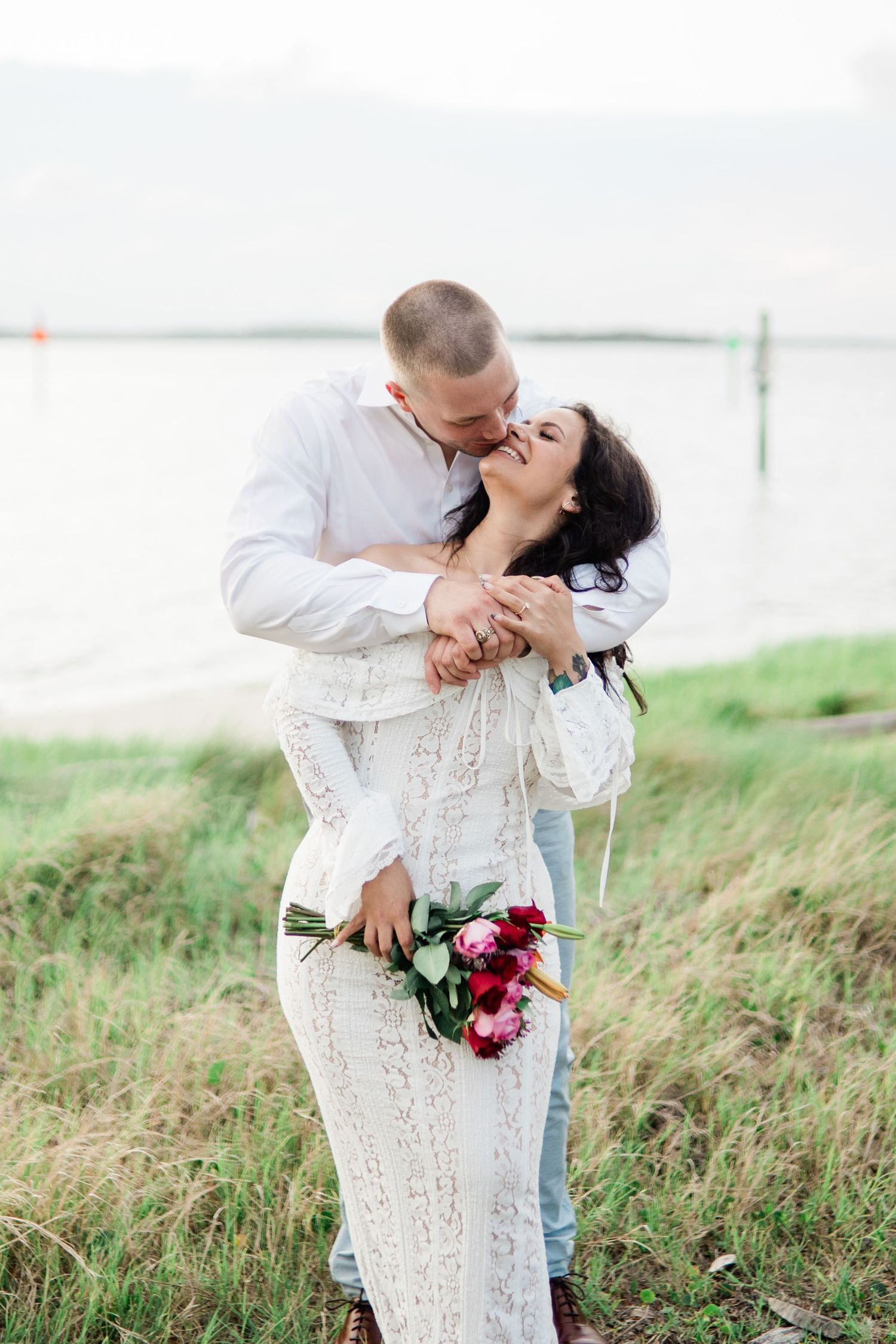 A romantic couple in white outfits share intimate moments while standing in a grassy field on their wedding day.