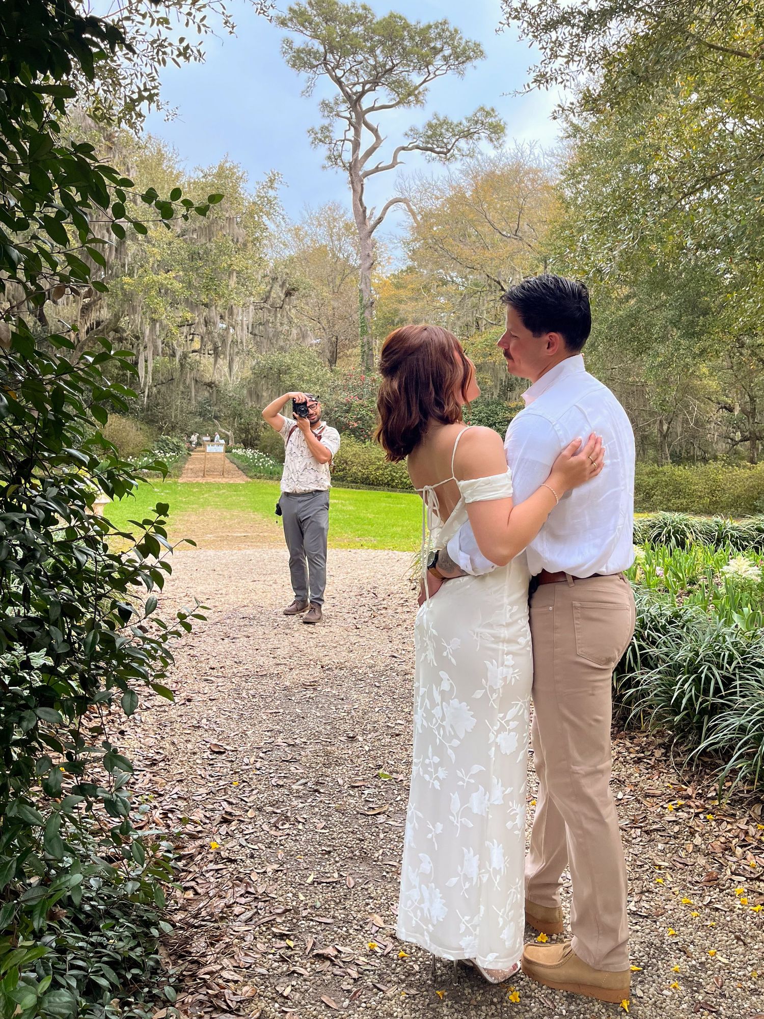 A photographer captures a couple in formal attire on a gravel path surrounded by lush garden scenery.