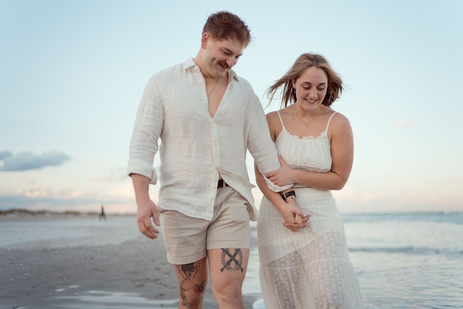 A couple in white attire walks along a beach at sunset while looking at their phones.