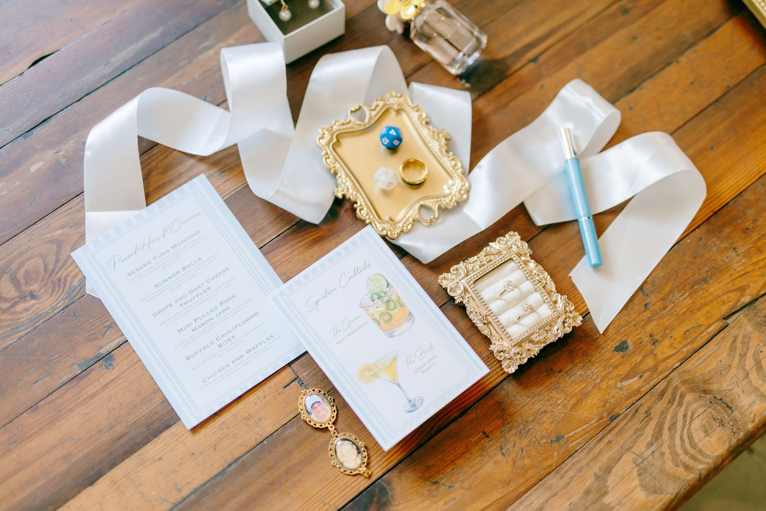 Elegant wedding flat lay with ivory invitations, gold ring box, ribbon, and jewelry on a rustic wood table.