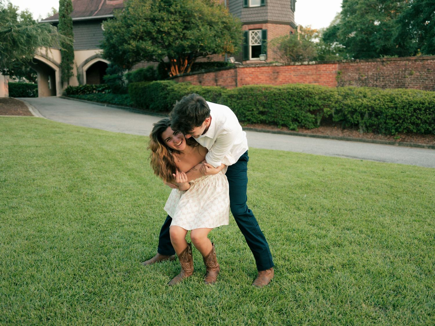 A couple embraces on a green lawn in front of a large brick house on a summer day.