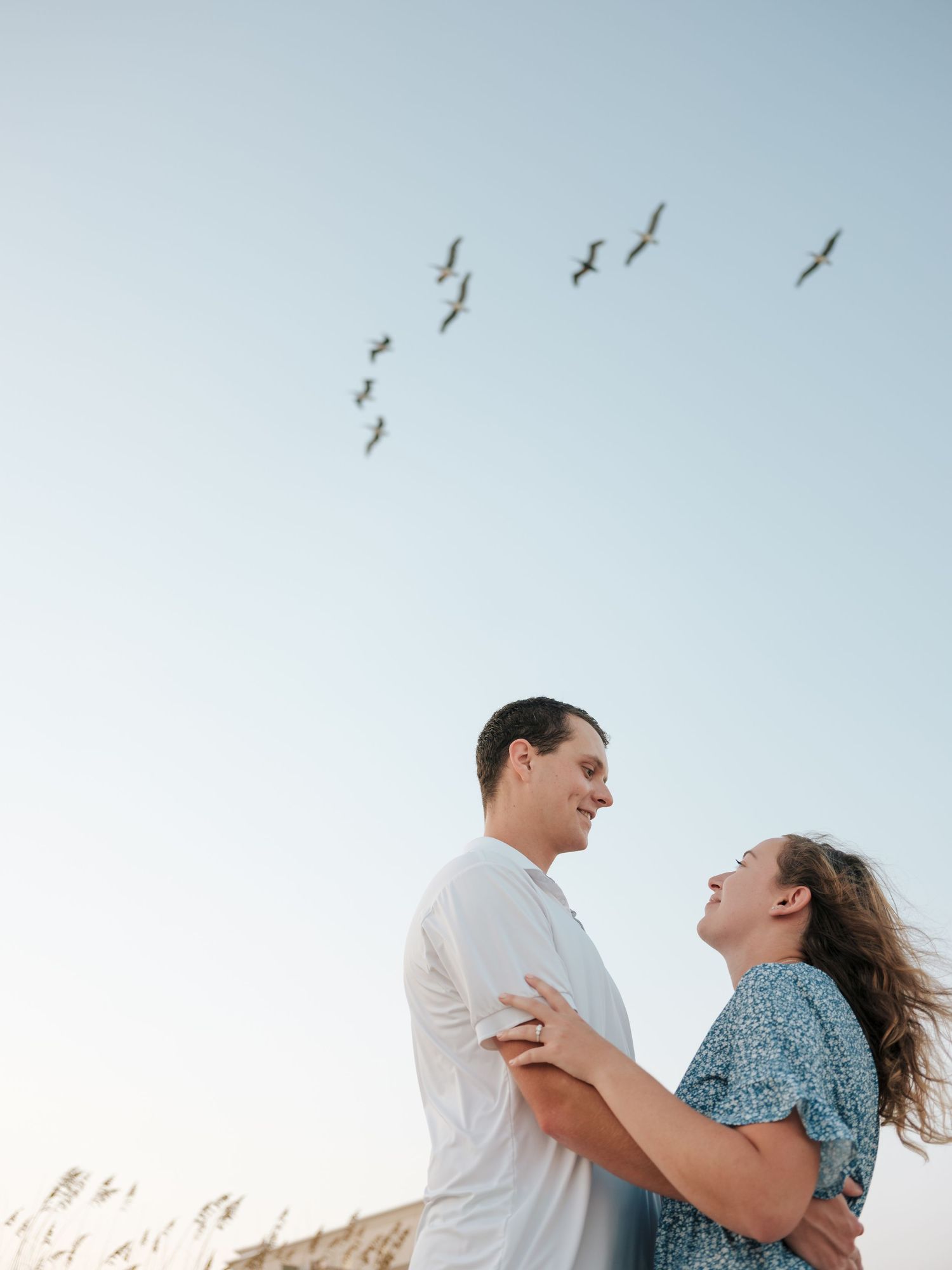 A couple embraces and laughs together on a sunny day with birds flying in the sky above.