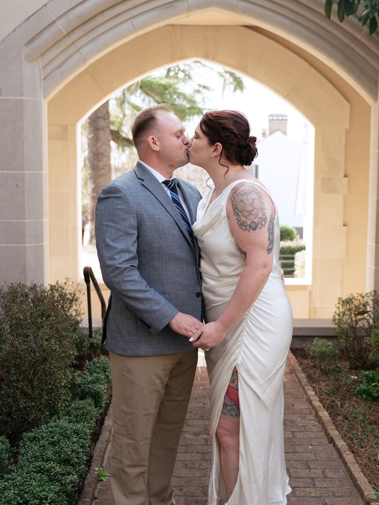 A romantic wedding kiss shared under a stone archway with greenery and landscaped gardens in the background.