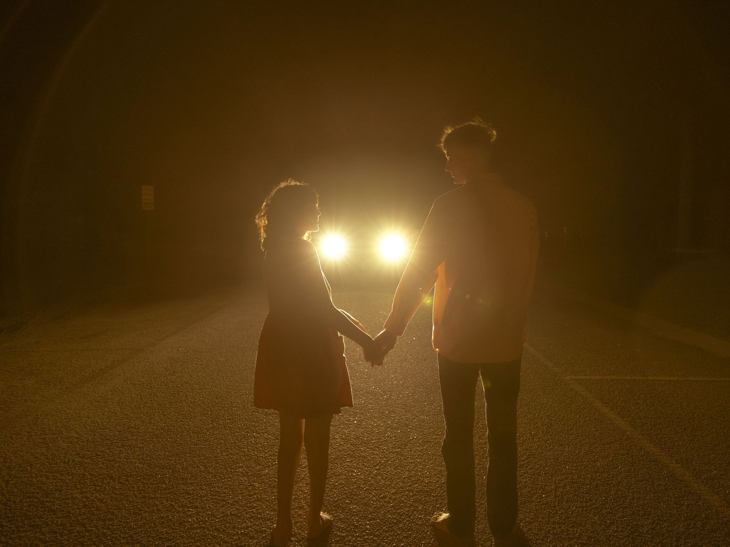 A row of silhouetted figures stand in darkness with car headlights glowing behind them on a foggy night.
