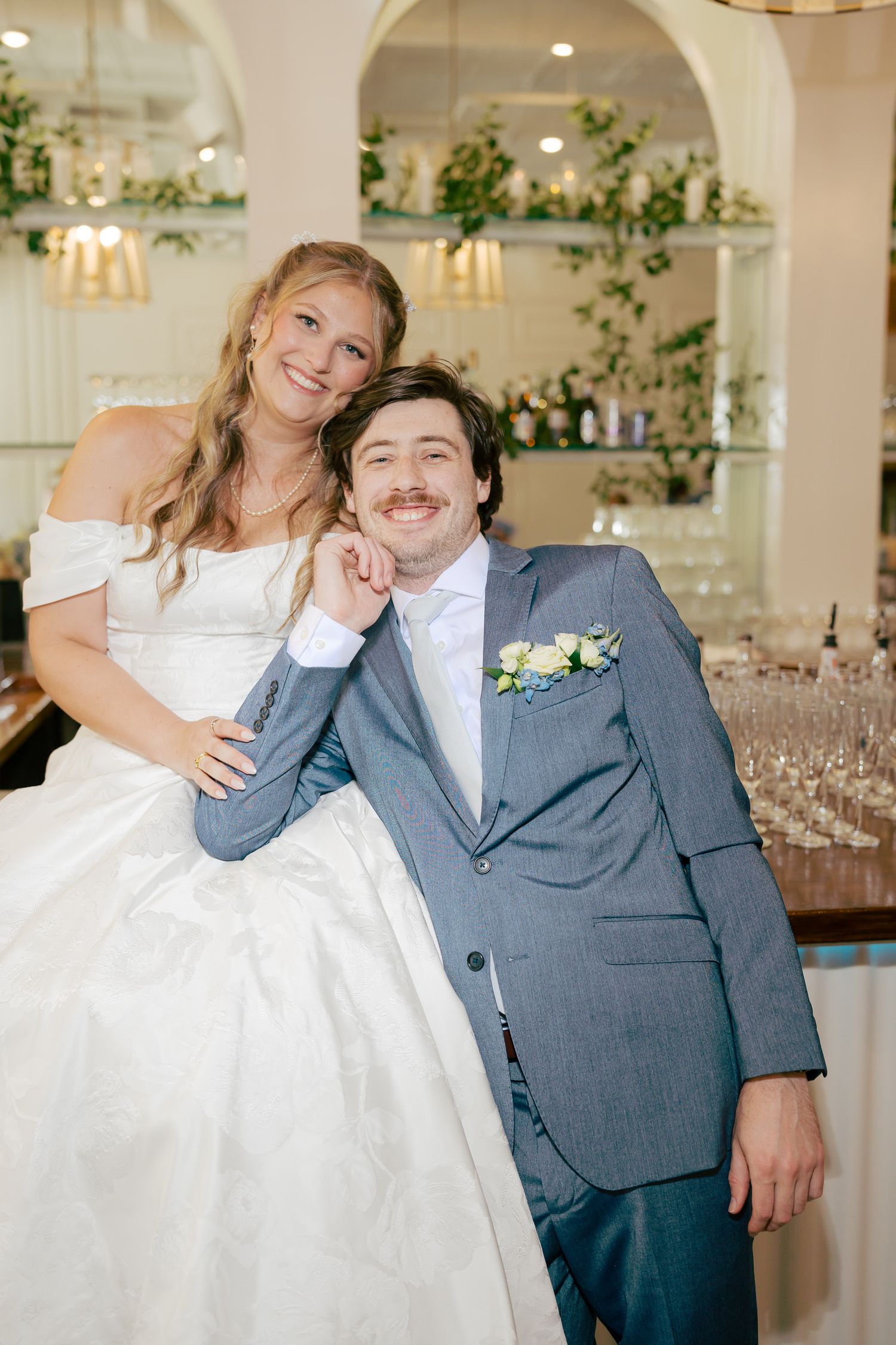 Happy bride and groom pose together at their wedding reception, she in a white gown, he in a gray suit with boutonniere.