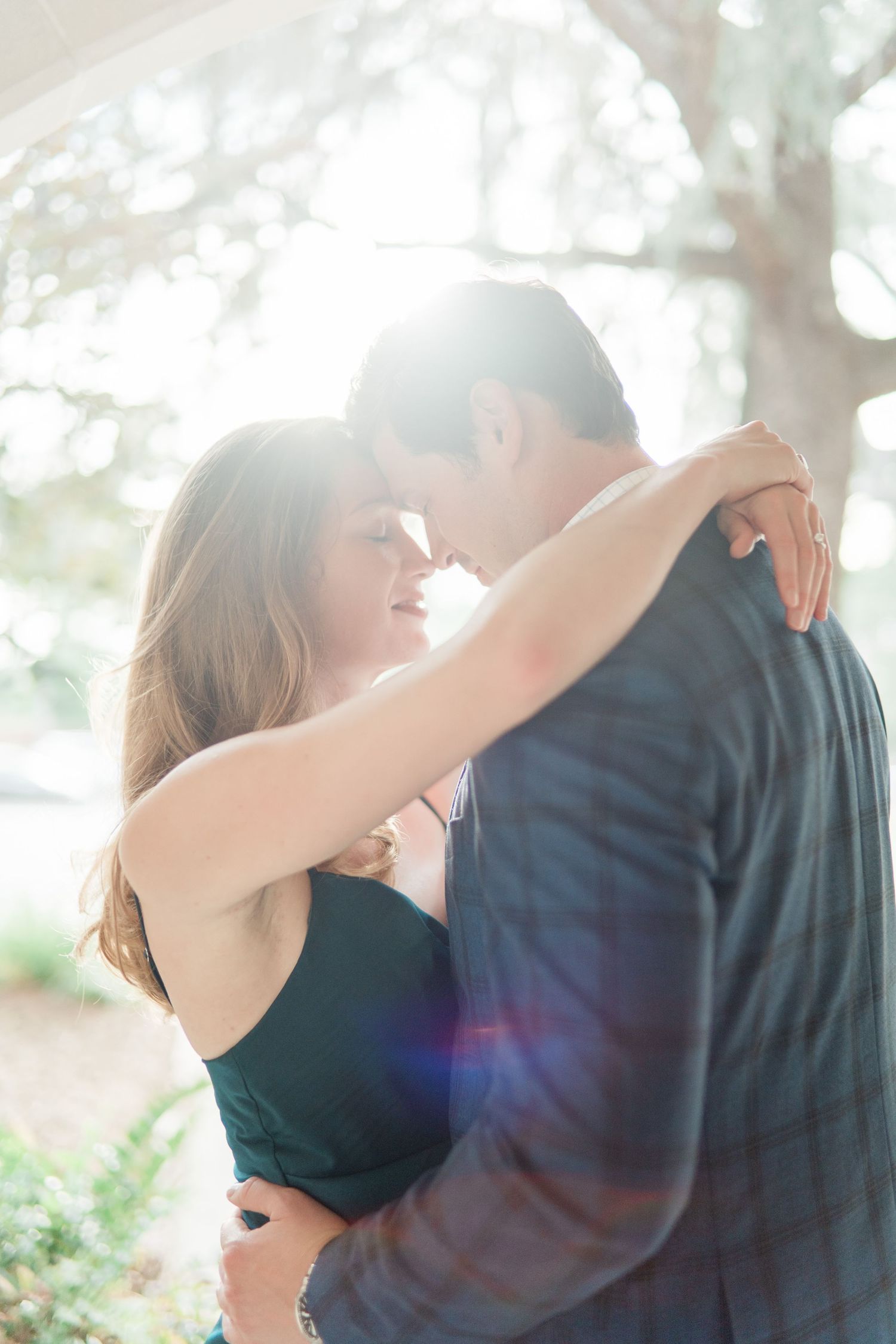 A romantic couple embraces in bright sunlight filtering through trees during an outdoor photo session.