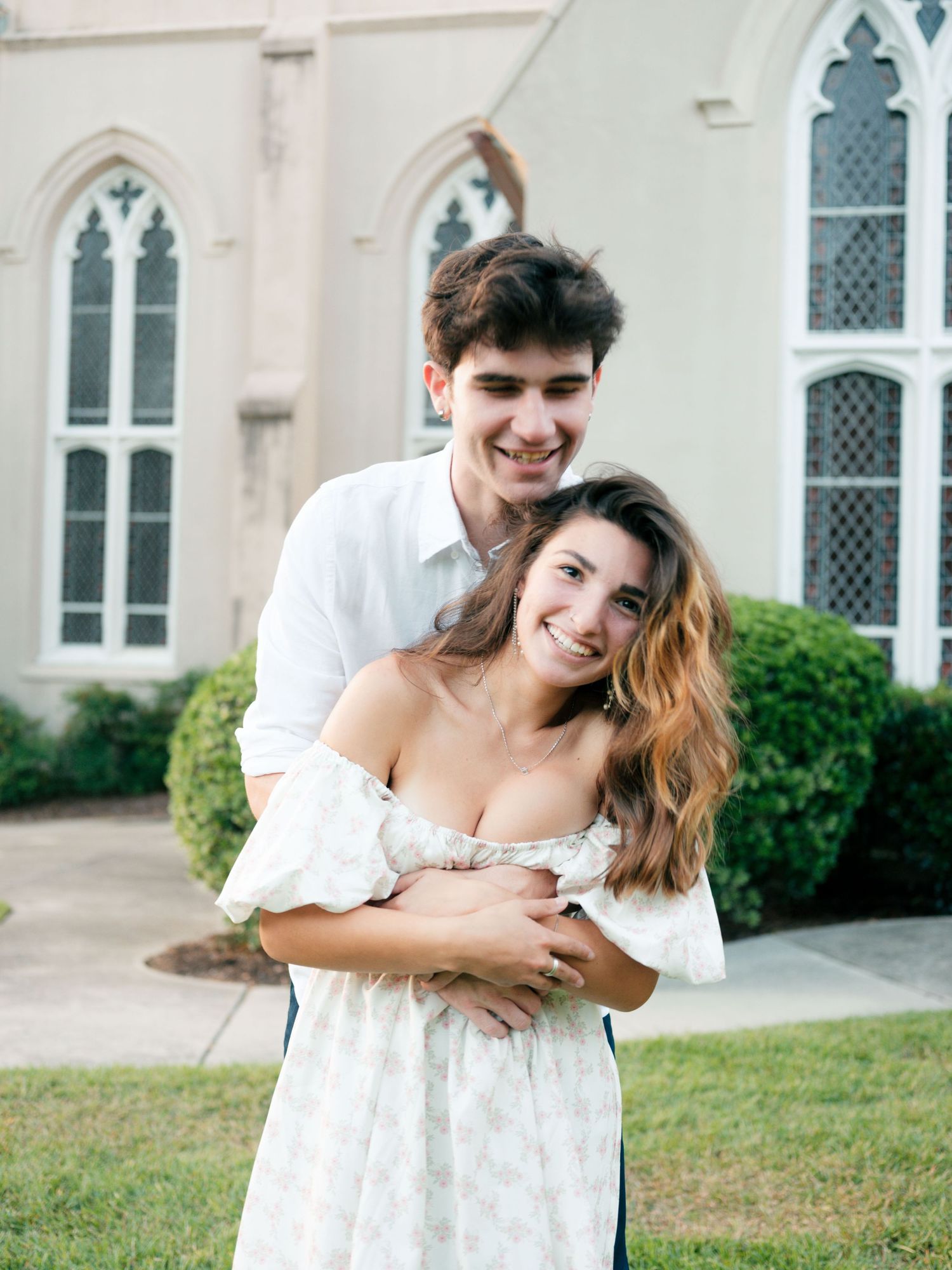 A romantic couple embraces while standing outside a Gothic church with arched windows on a sunny day.