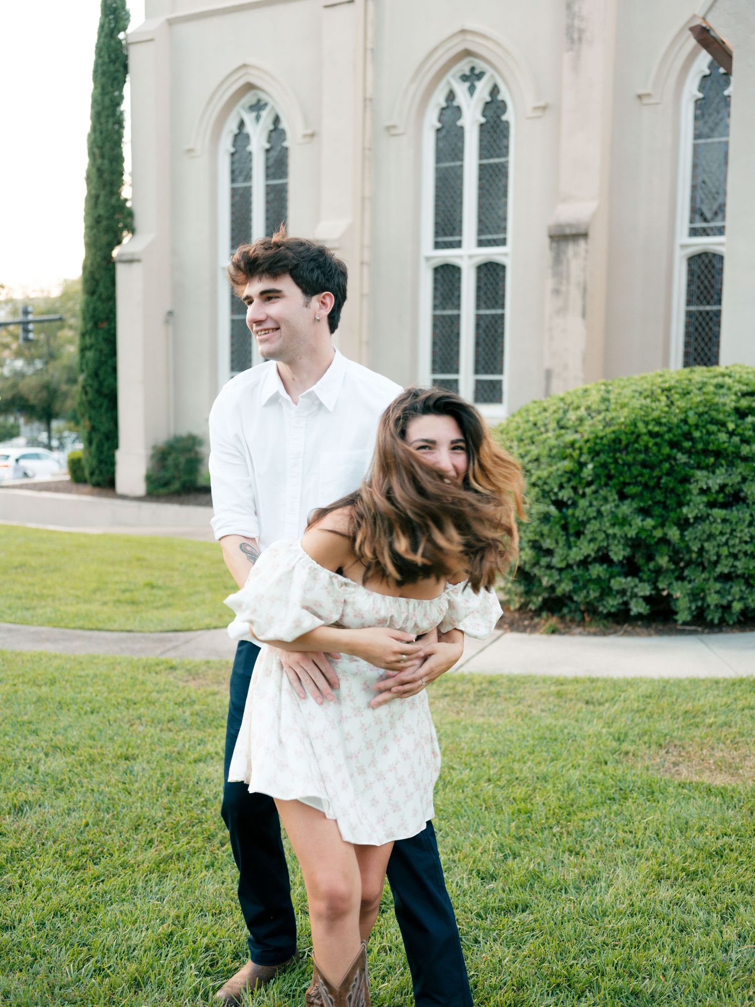 A romantic couple embracing on a lawn in front of a Gothic-style church with tall arched windows.