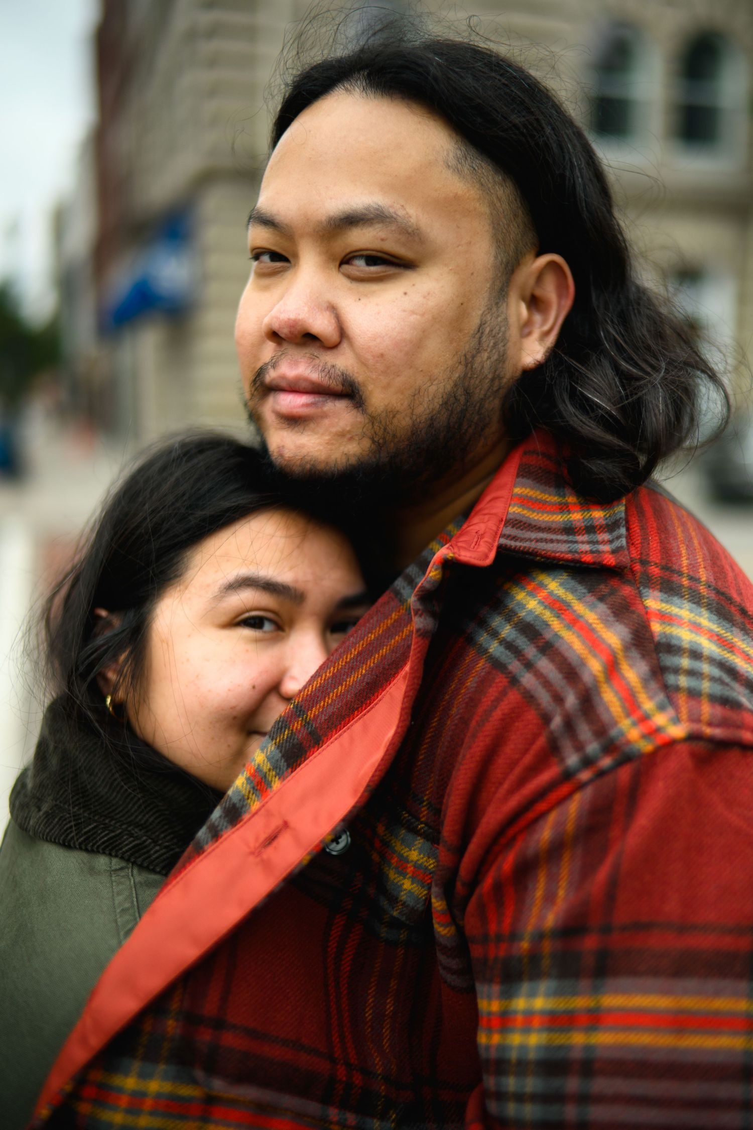 A couple embraces while wearing winter clothing, with one in a red plaid flannel jacket standing against an urban backdrop.