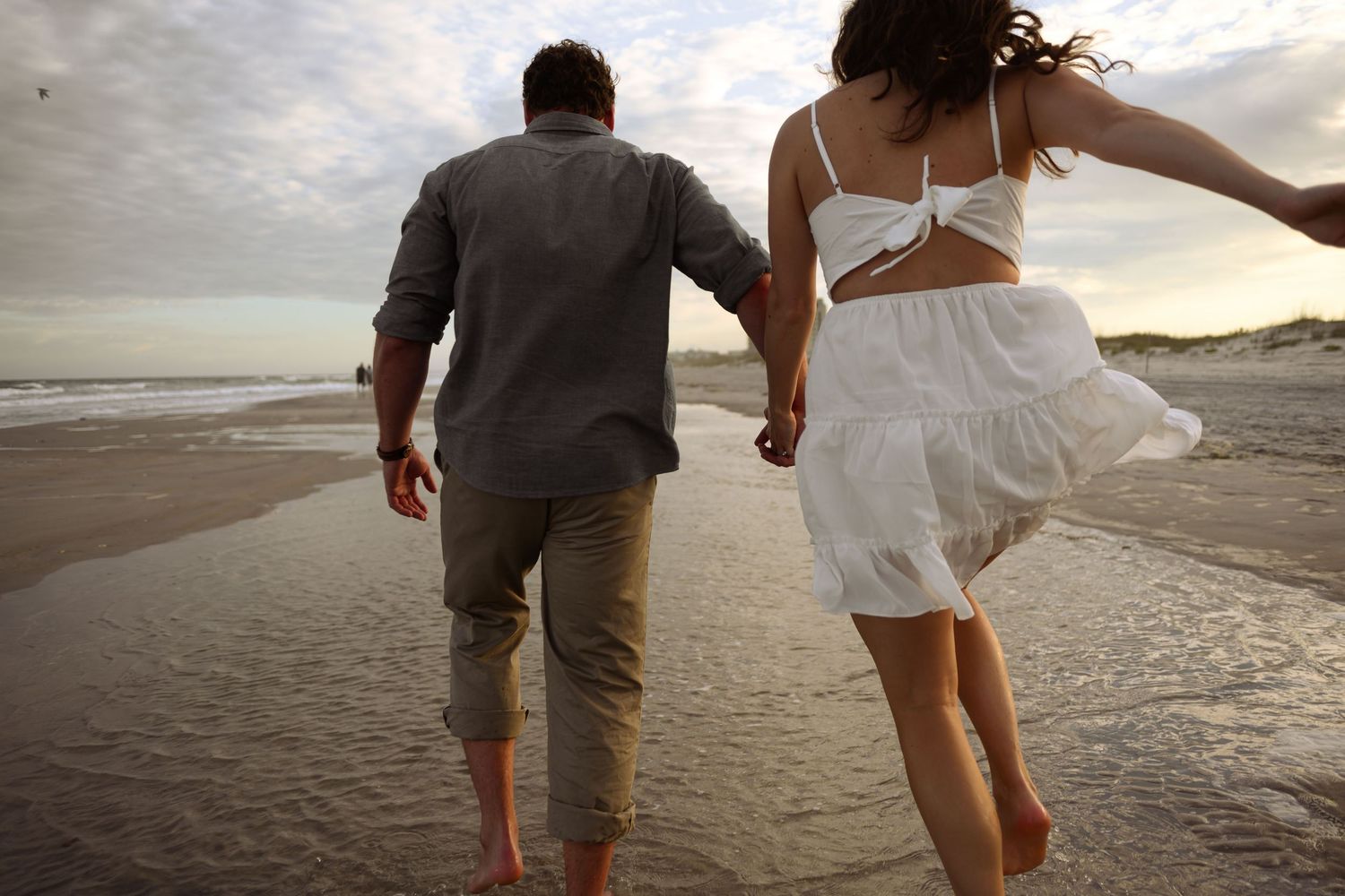 A couple walks hand in hand along a beach shoreline at sunset with waves gently rolling in.