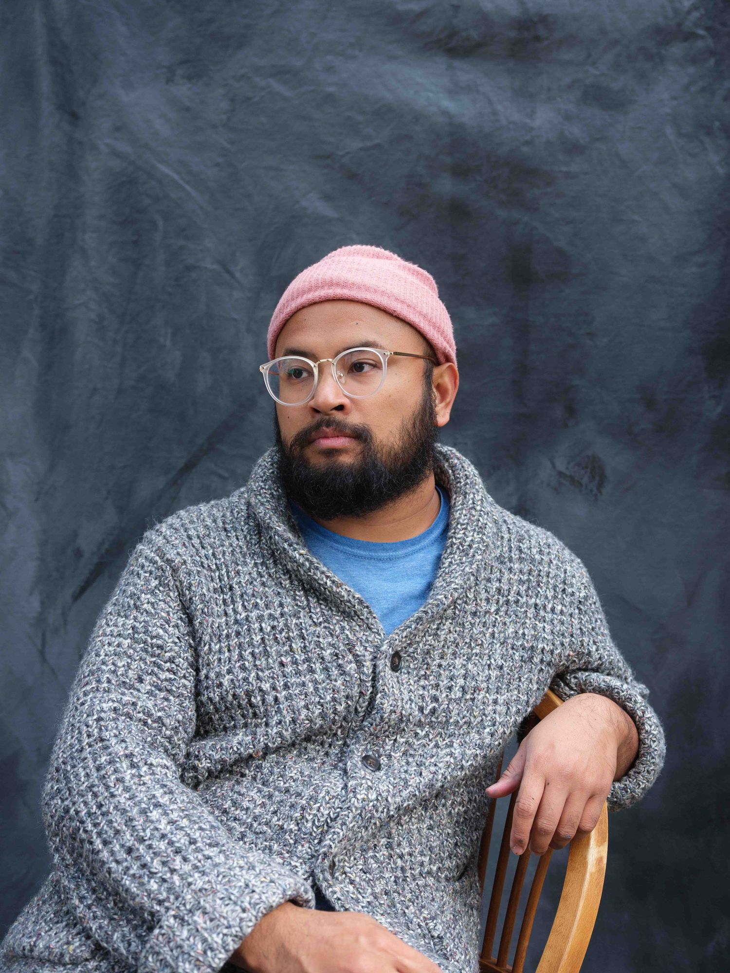 Portrait of someone wearing a pink beanie, glasses and grey knit cardigan seated against a dark background.