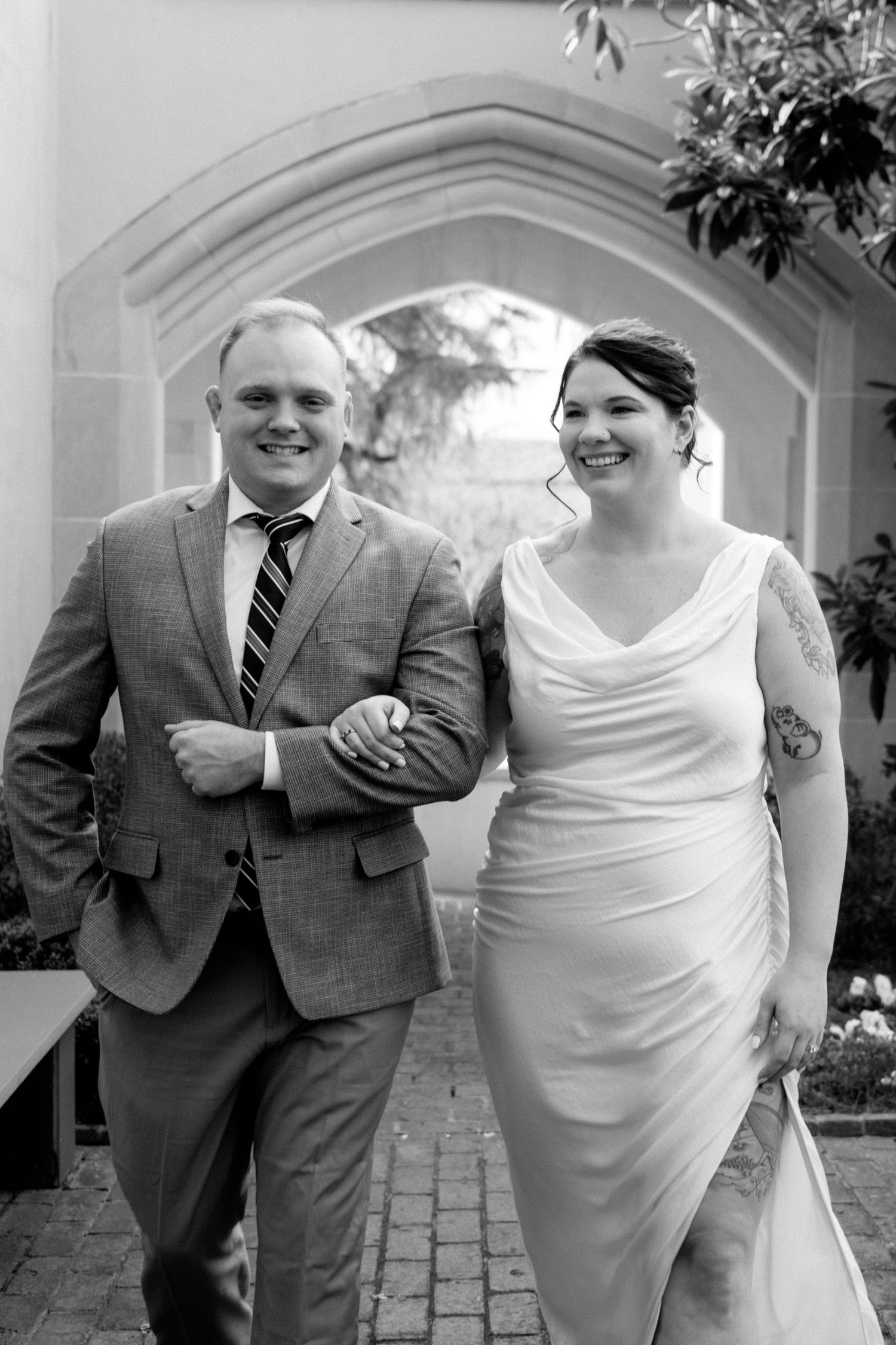 A joyful formal couple walking arm-in-arm through an arched courtyard in black and white wedding attire.