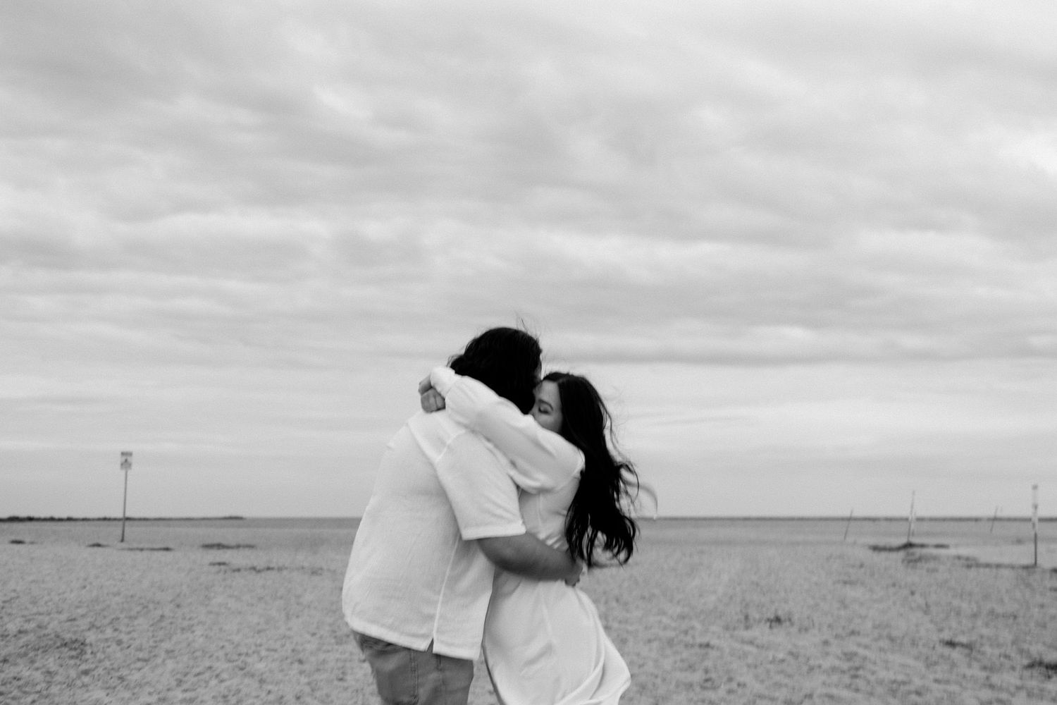 A black and white photo series showing a couple embracing on a beach with sailboats in the background.
