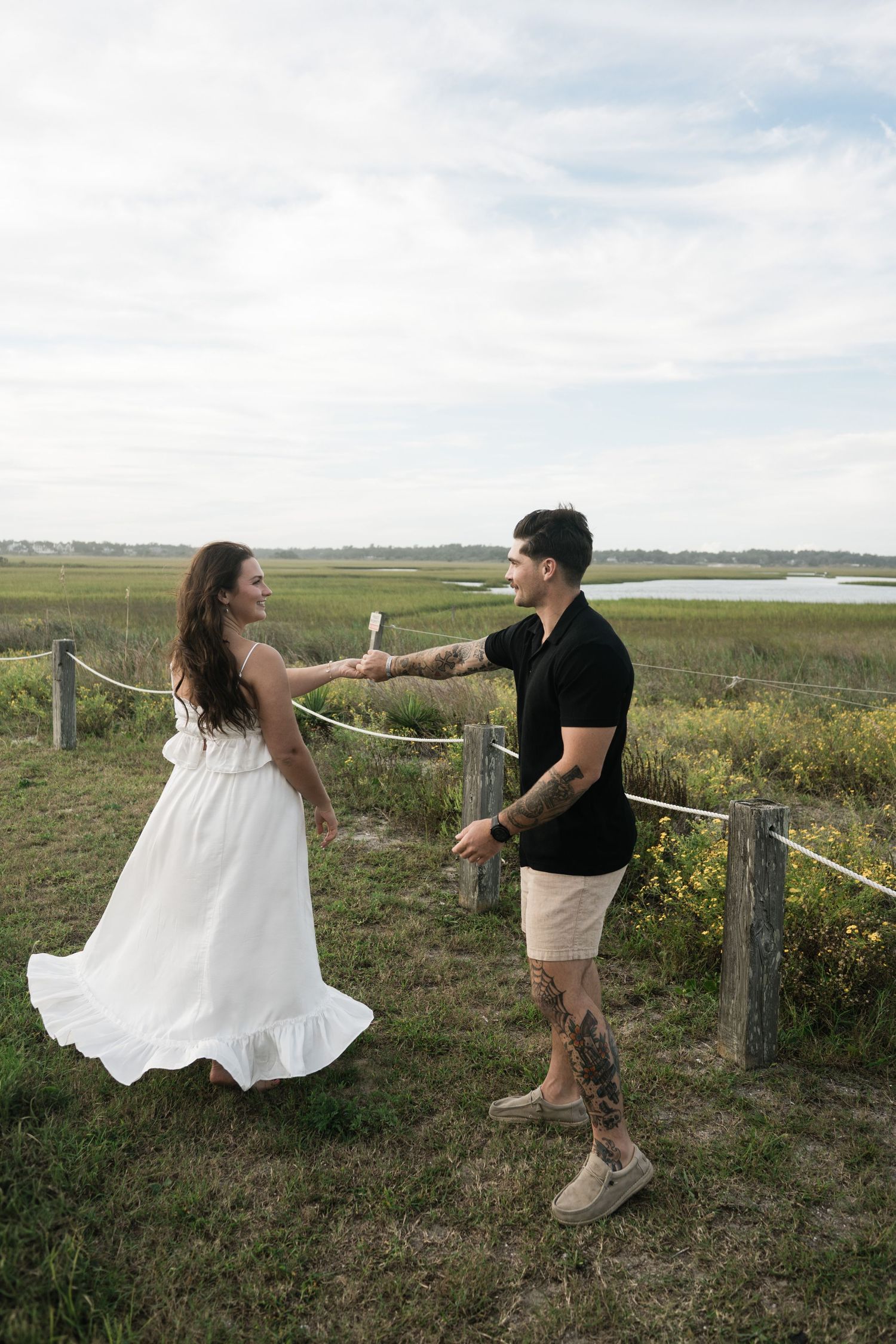 A couple in casual attire dances playfully together on grassy coastal terrain with a fence in the background.